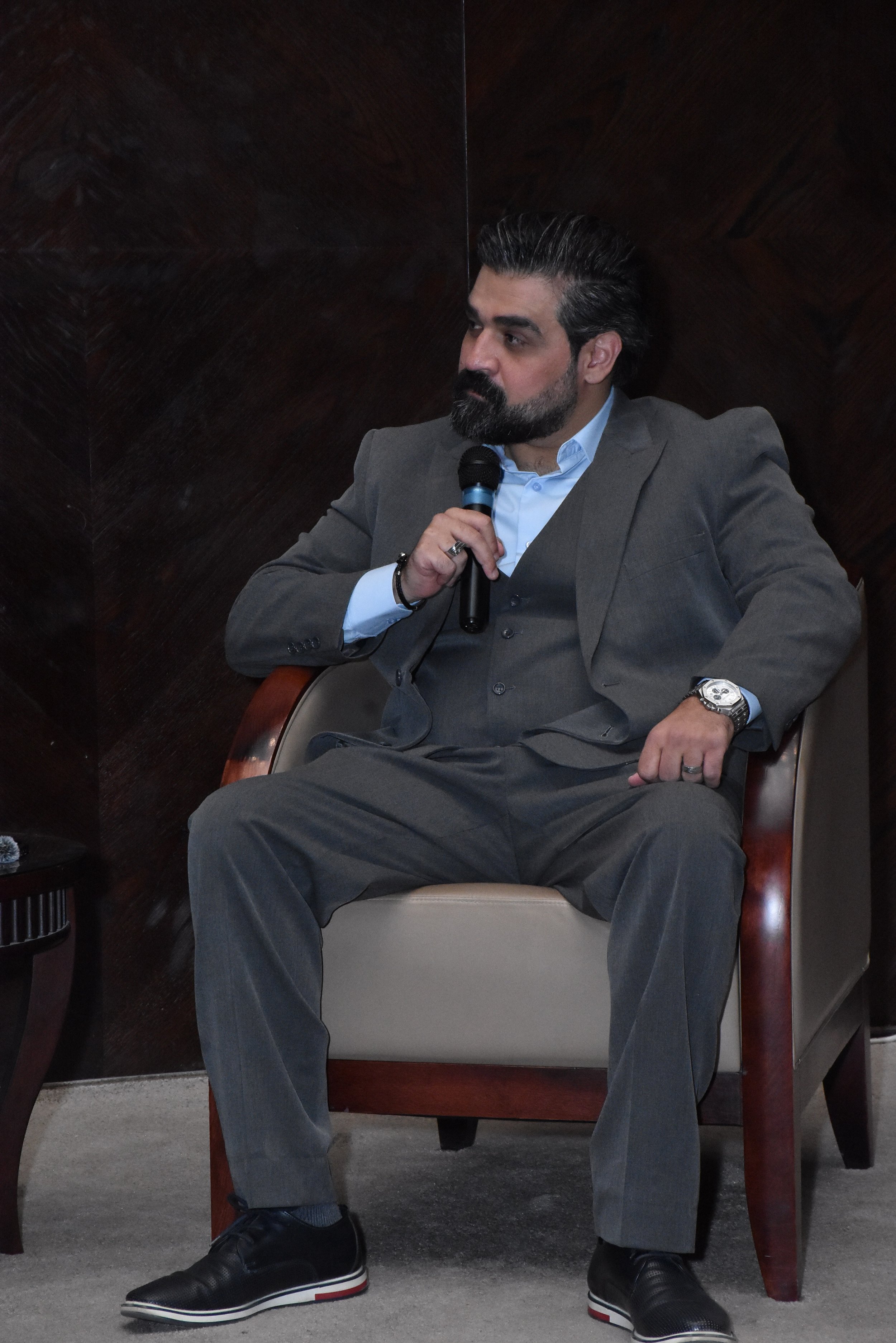 A man in a gray suit sitting in a beige armchair, holding a microphone, with dark hair and beard, in an indoor setting.