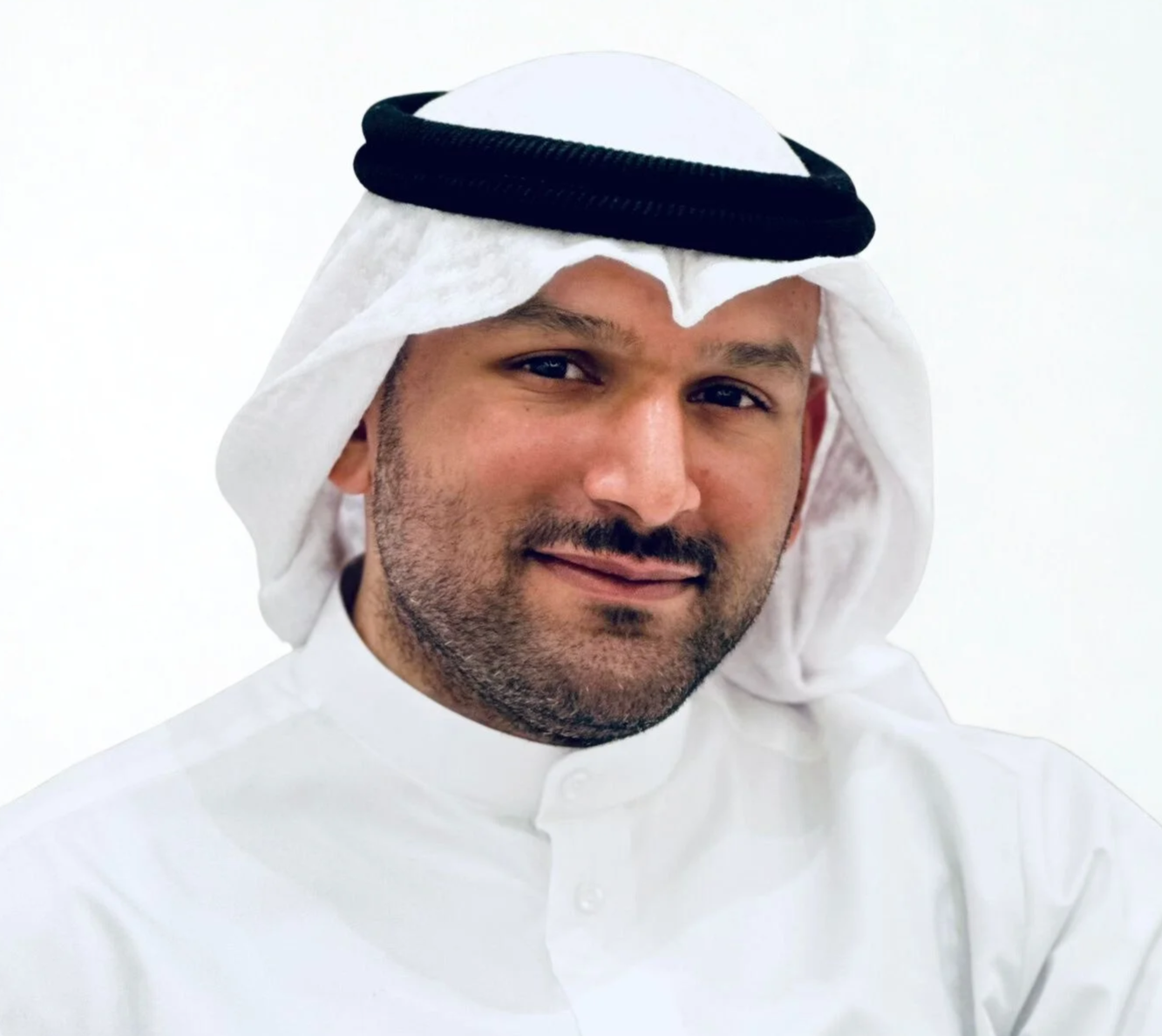 A man wearing traditional Middle Eastern attire, including a white kandura and a white ghutra with a black agal, smiling against a plain white background.