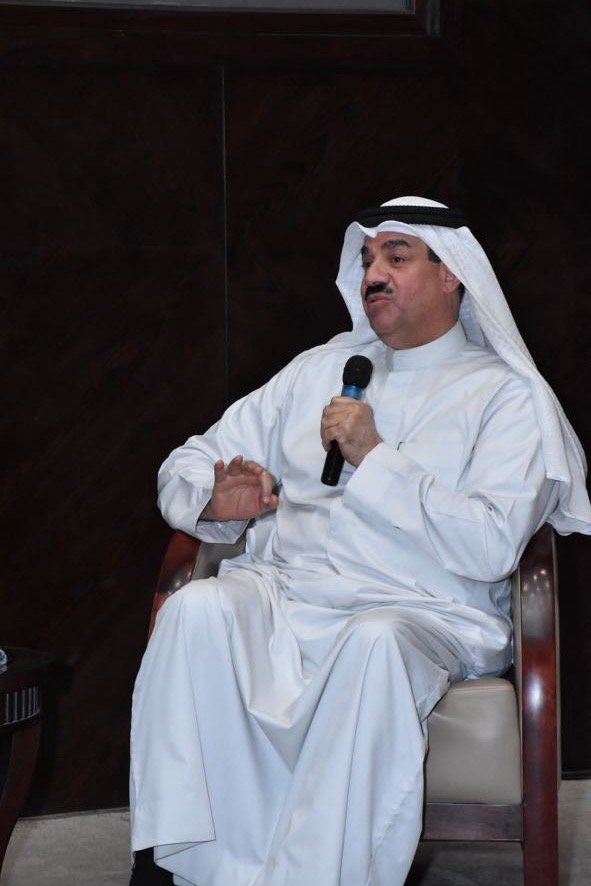 A man dressed in traditional Middle Eastern attire, including a white thobe and a keffiyeh, is sitting on a chair and speaking into a microphone.