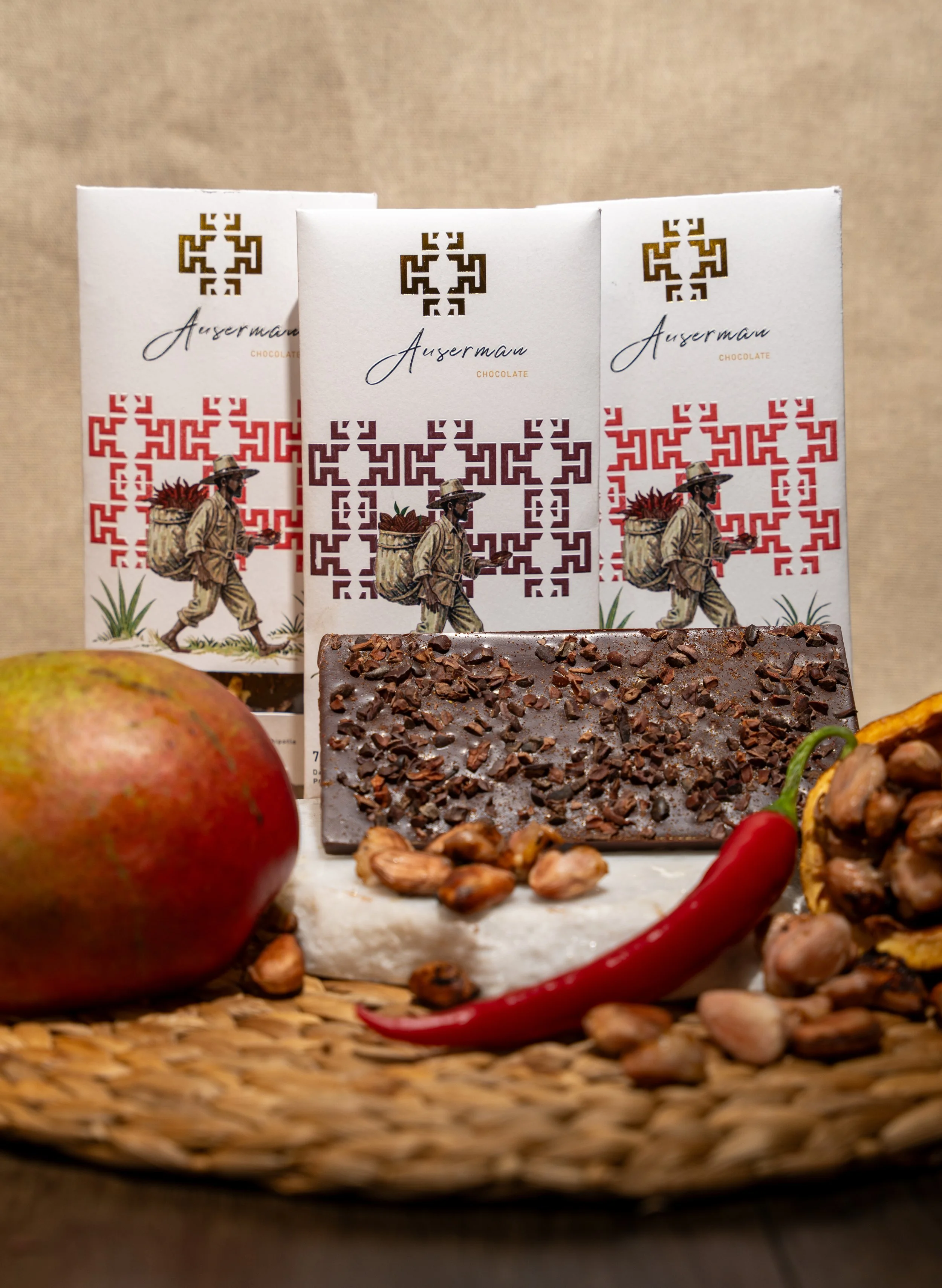 Three bars of dark chocolate with chopped nuts on top, apple, red chili, and raw nuts arranged on a woven tray in front of chocolate packaging with a man walking image.