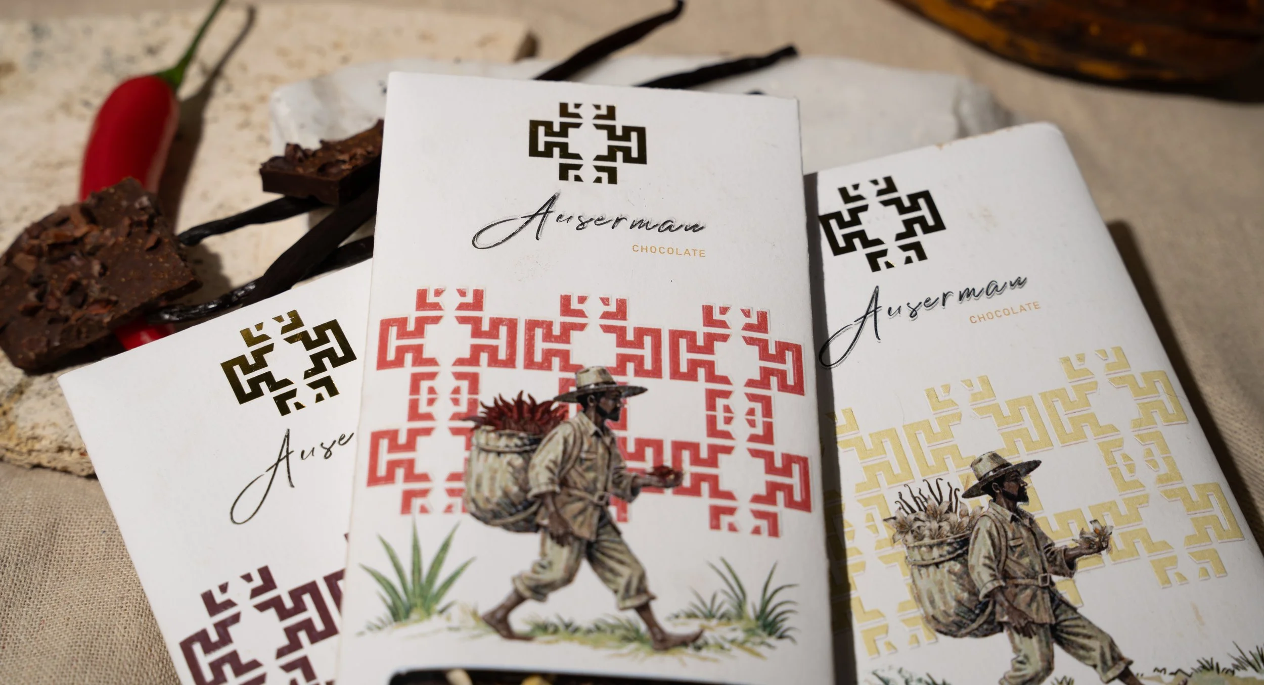 Three boxes of Auserman chocolate with a vintage illustration of a man with a backpack and a walking stick, set on a textured surface with a chili pepper and chocolate pieces nearby.