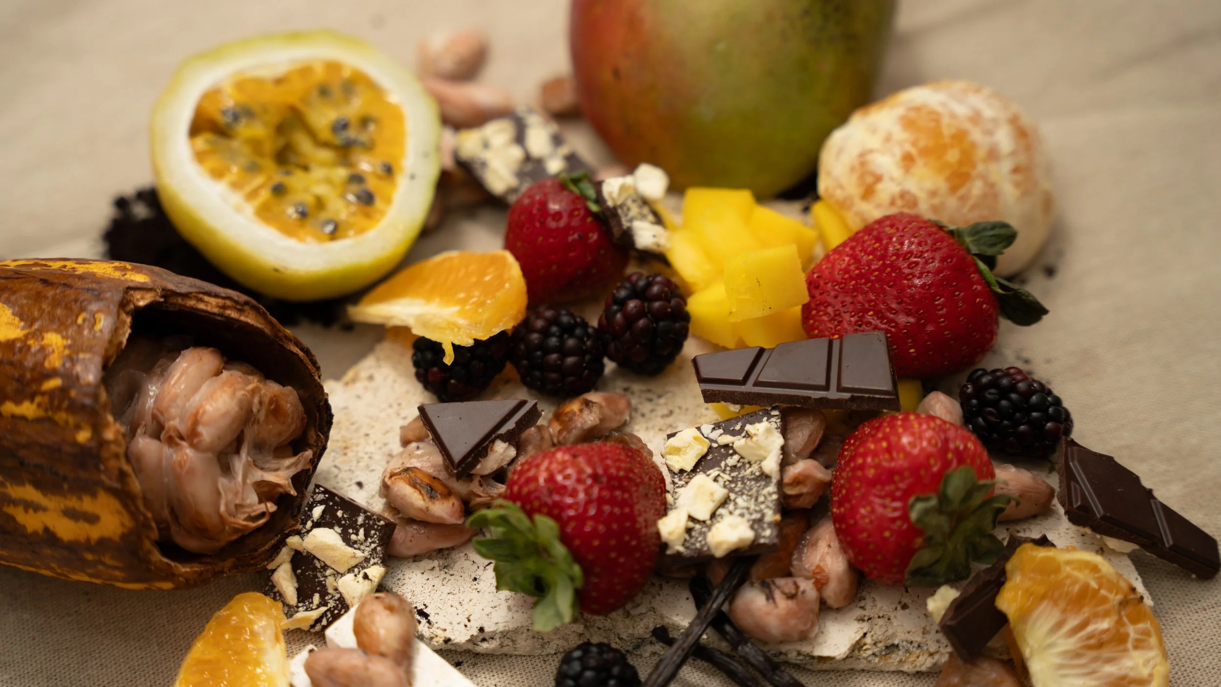 Assorted fruits such as strawberries, blackberries, orange slices, passionfruit half, apple, and mango pieces, along with chocolate pieces and nuts on a surface.