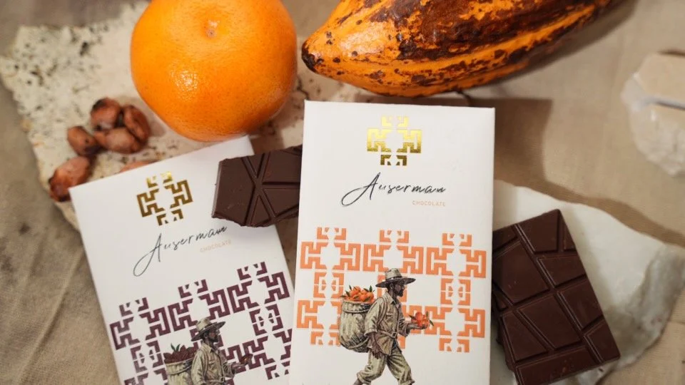 Two bars of dark chocolate, an orange, a banana, a few bitter nuts, and a piece of clear quartz on a beige surface.