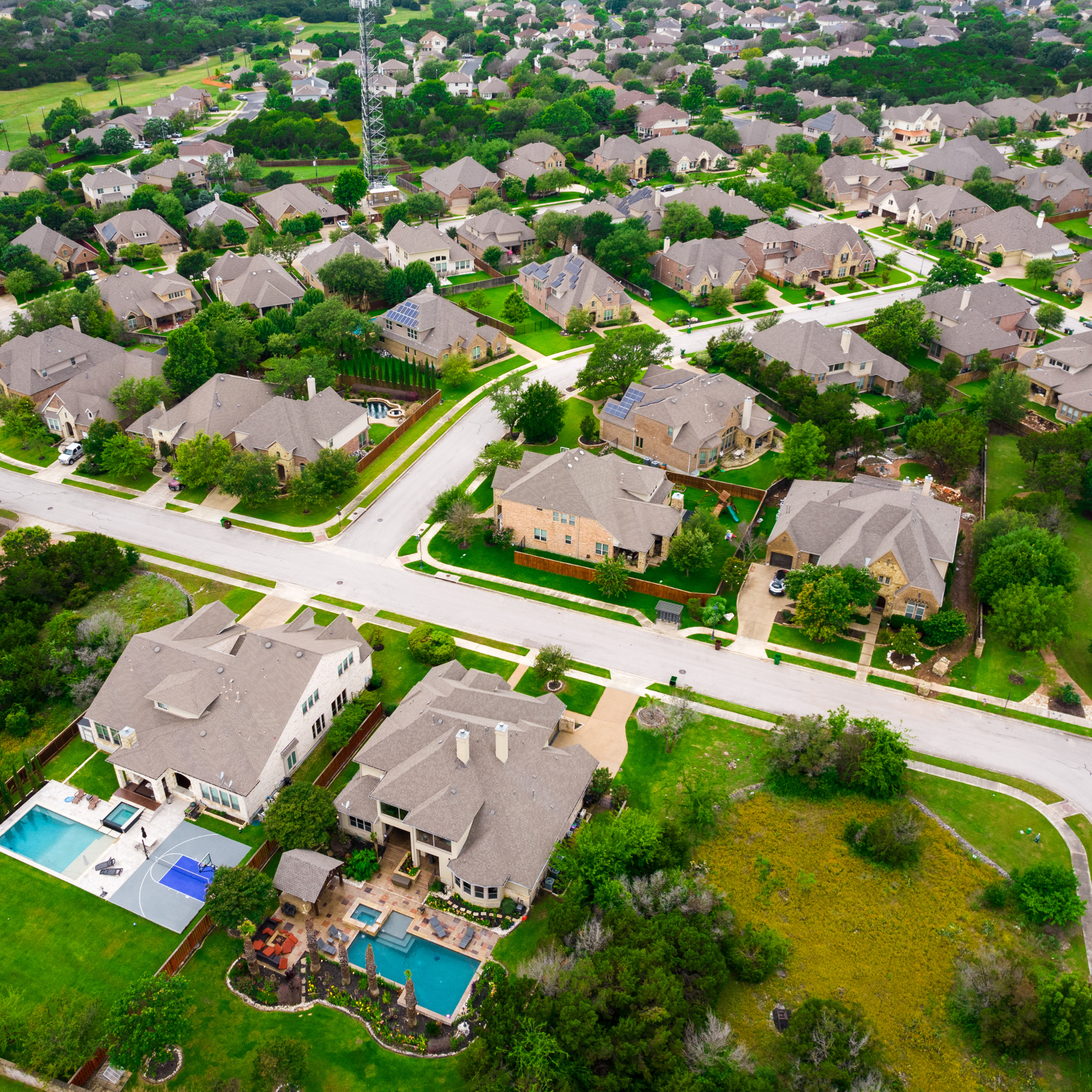 Aerial view of a suburban neighborhood with numerous single-family homes, green lawns, trees, and streets. Several houses feature swimming pools and solar panels.