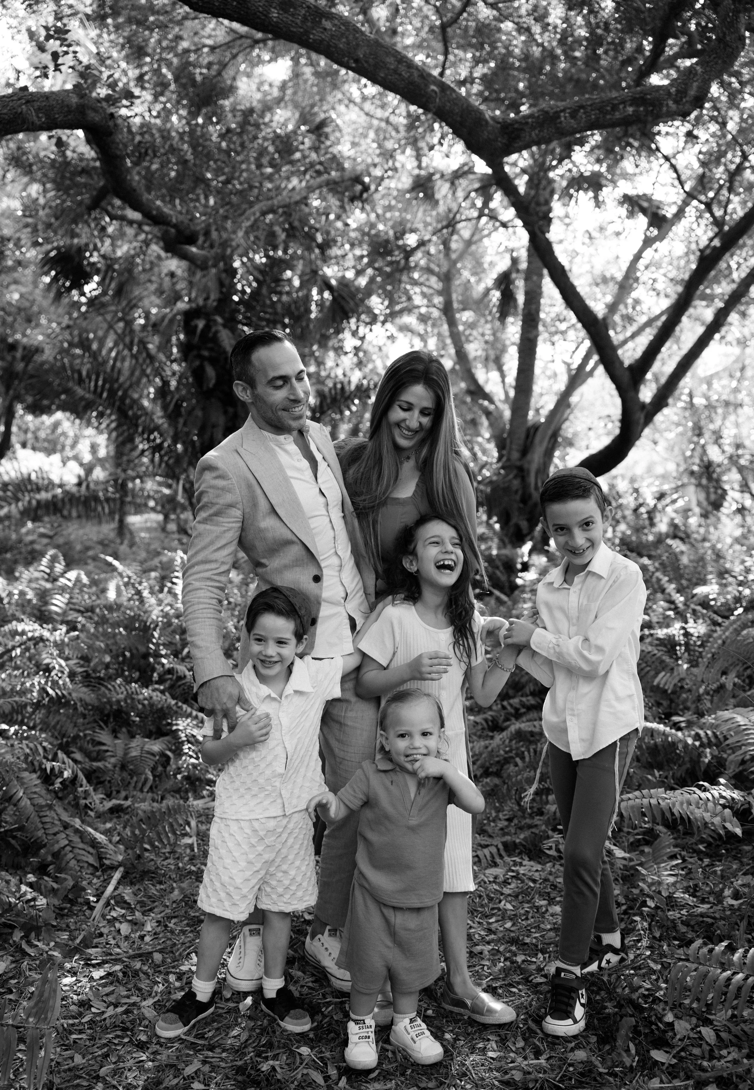 A family of six, including two adults and four children, smiling and laughing together outdoors in a lush, wooded area.