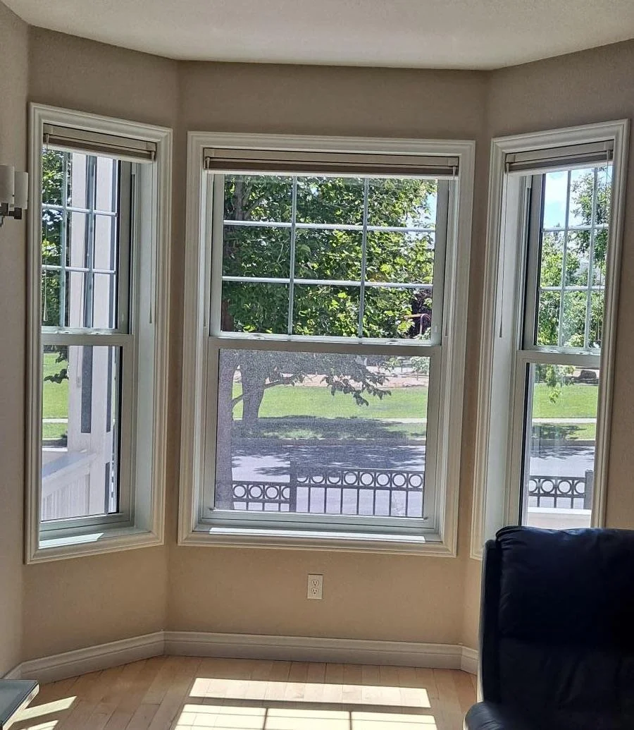 A bright room with three large windows showing green trees outside and a street. The windows have white trim and are partly covered with blinds. There is a black chair in the lower right corner and a white wall with an electrical outlet.