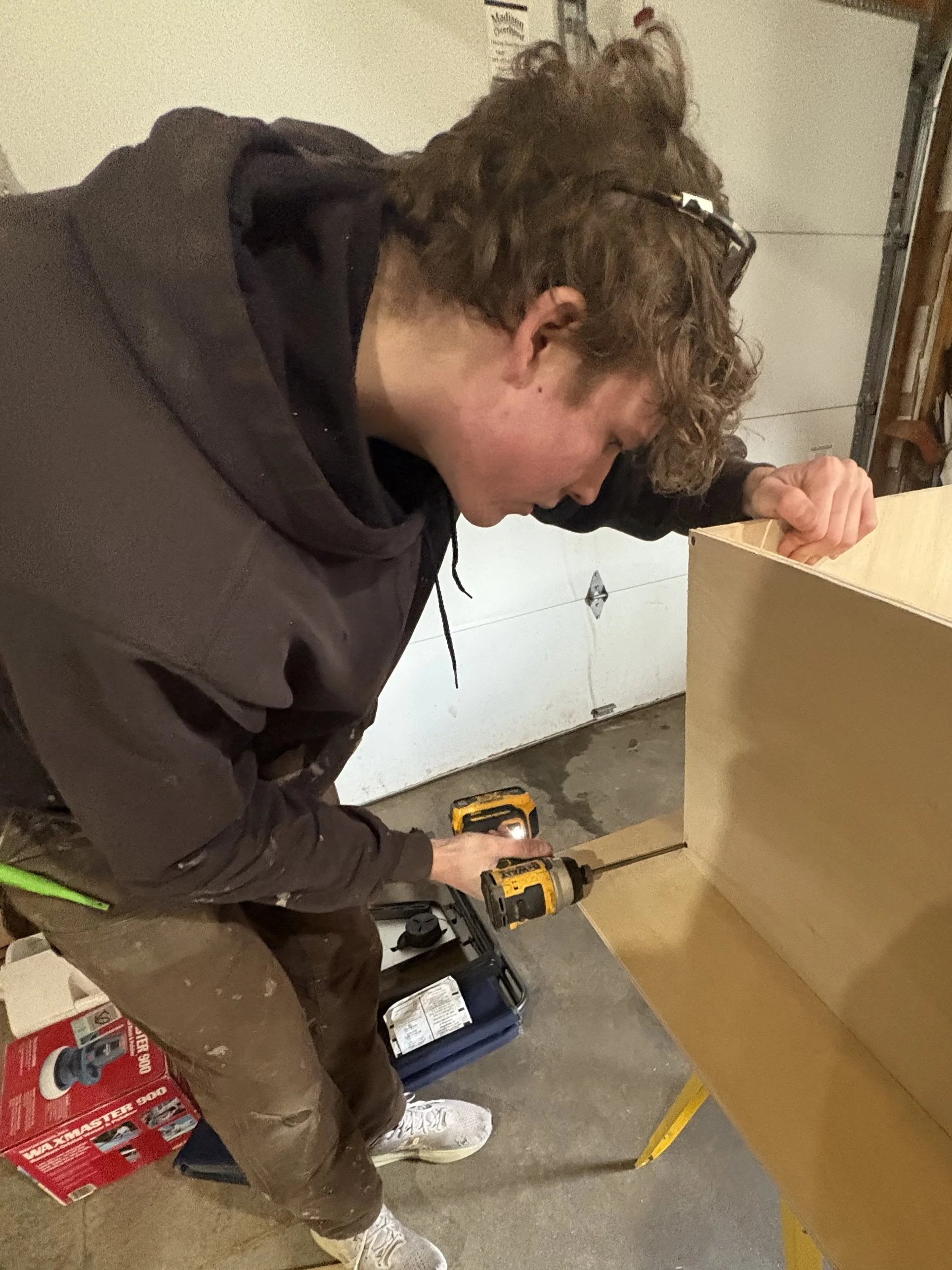 A person with curly brown hair, glasses, wearing a black hoodie and brown pants, is using a yellow power drill on a wooden piece, working on a DIY project in a garage or workshop setting.