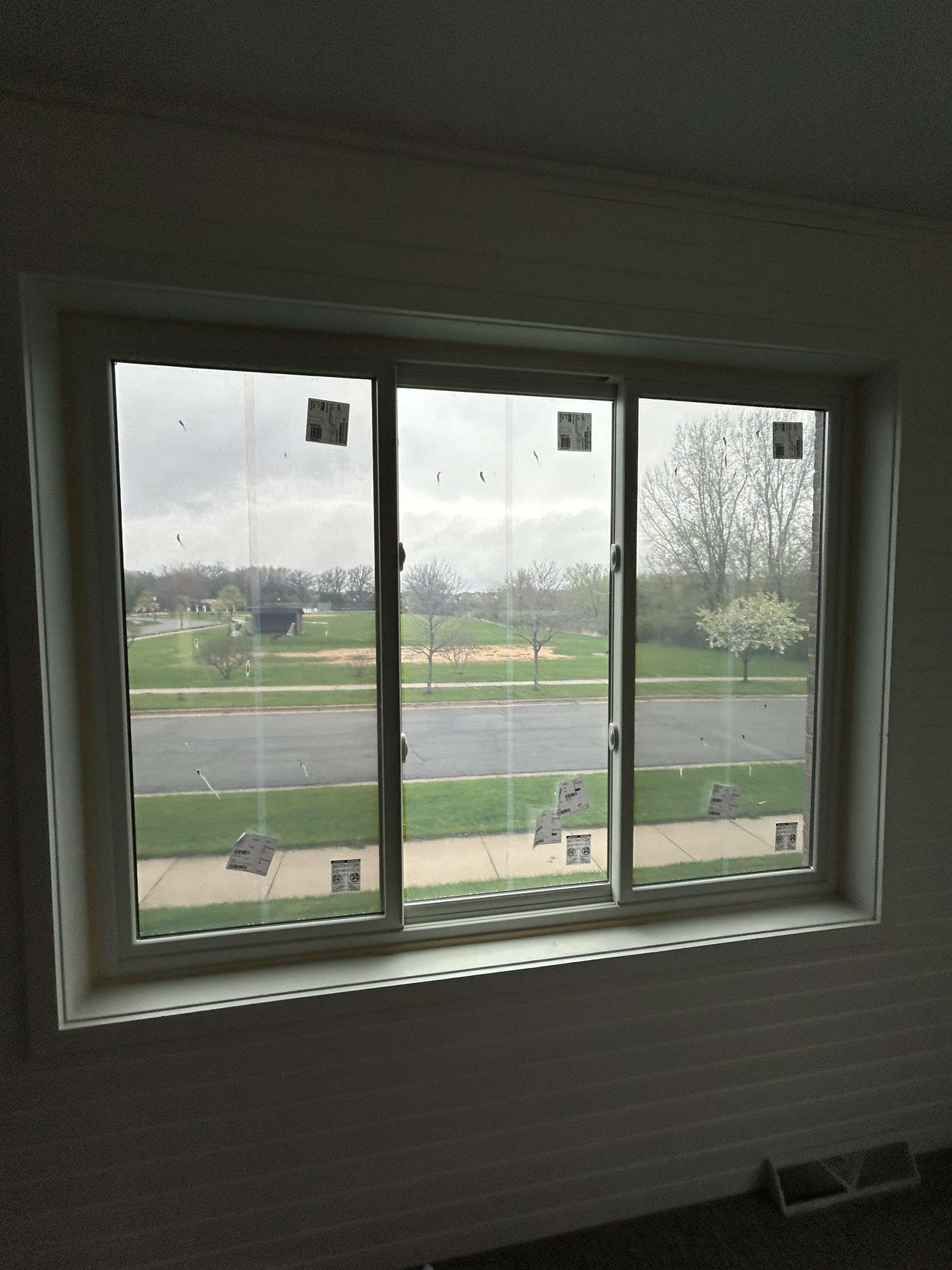 A view through a large window showing a street, grassy area, trees, and cloudy sky outside.