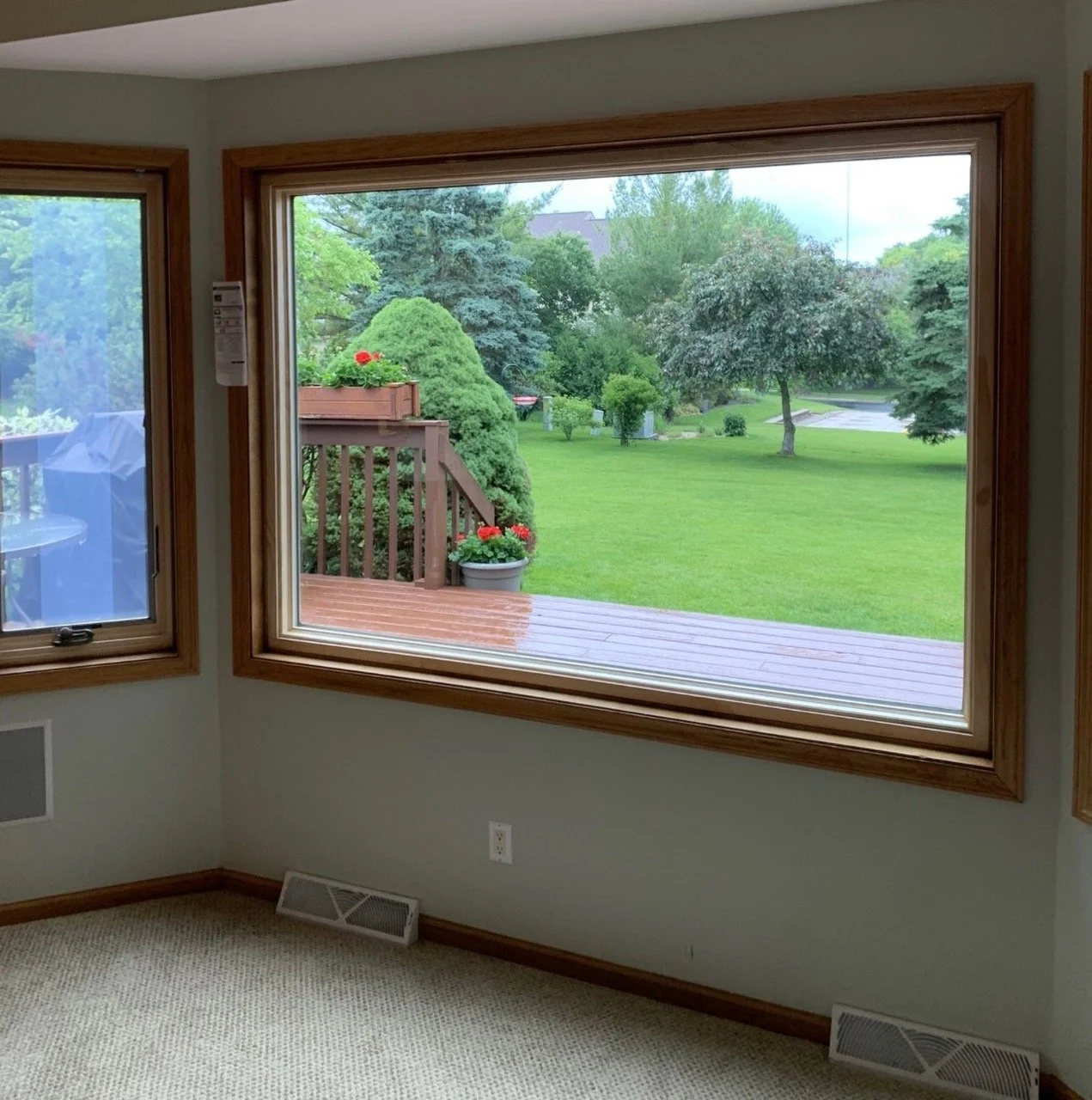View through large window showing a backyard with a wooden deck, potted plants with red flowers, trimmed bushes, and trees with green foliage.