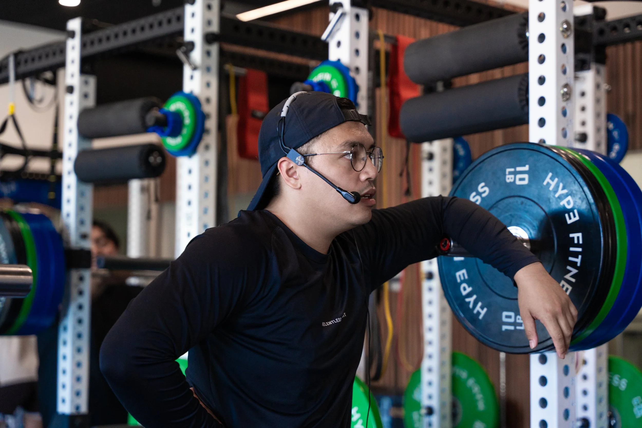 A man wearing glasses, a black cap backwards, and a headset with microphone is in a gym, lifting a barbell with blue weight plates labeled 'HYBRID FITNESS 10 LB'. The background shows gym equipment and other people.