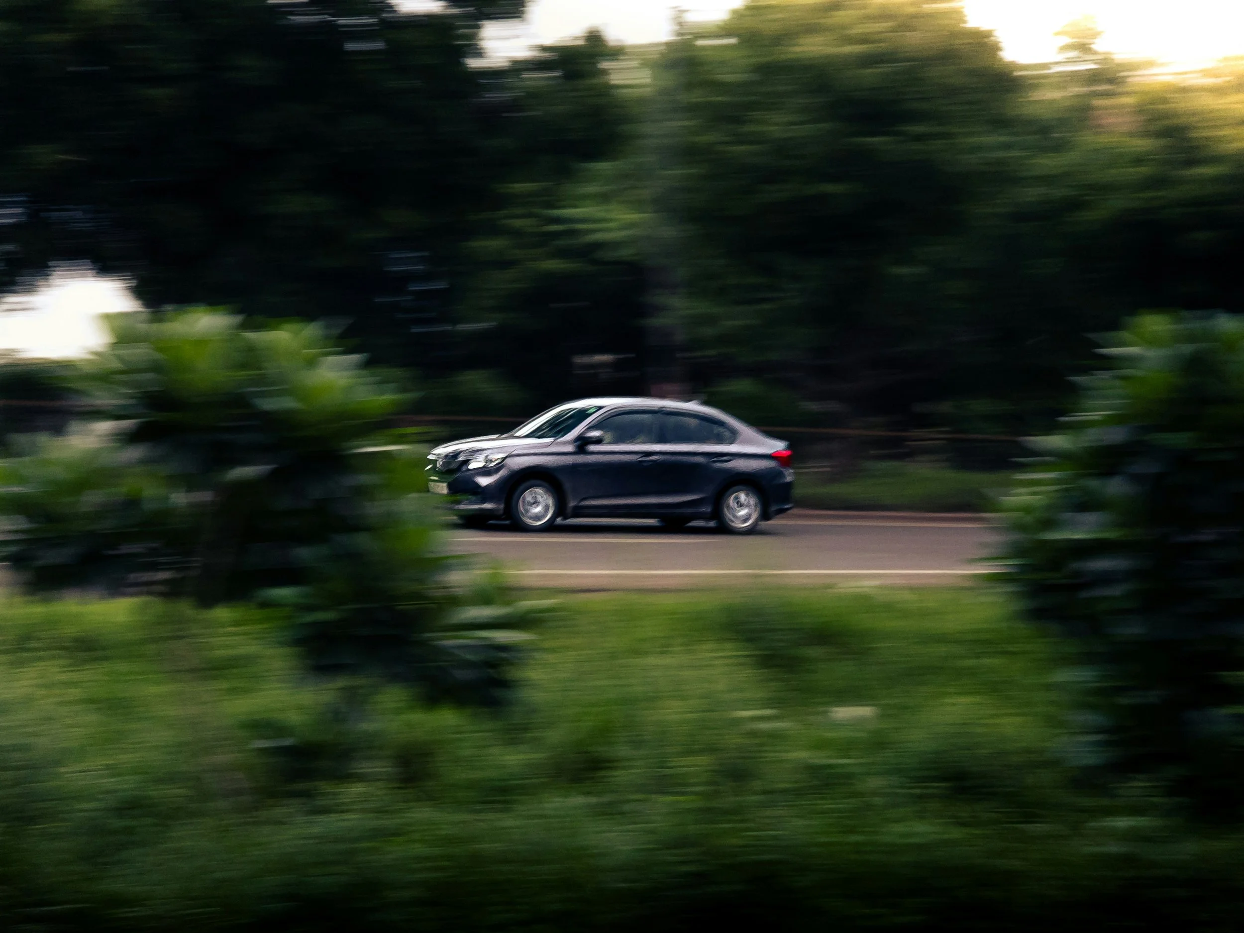 Schwarzfahrender Pkw auf einer Straße, umgeben von grünen Büschen und Bäumen, bei Sonnenuntergang