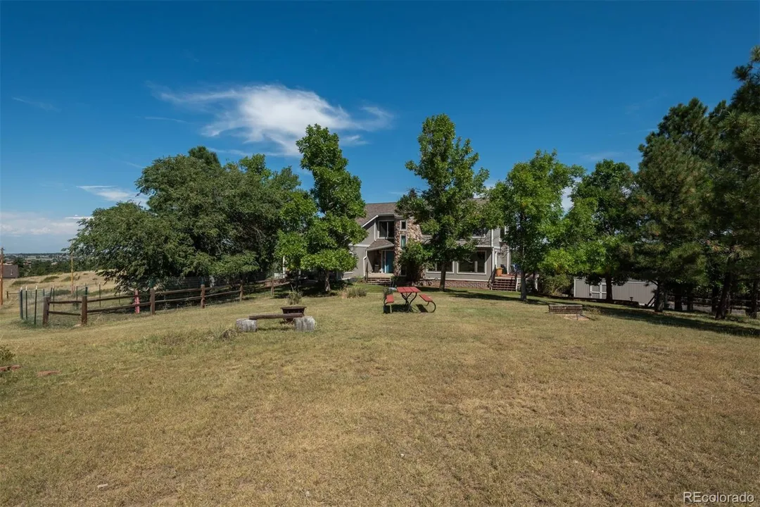 Large backyard with a house in the background, surrounded by trees, under a bright blue sky with some clouds, with outdoor furniture including benches and a table.