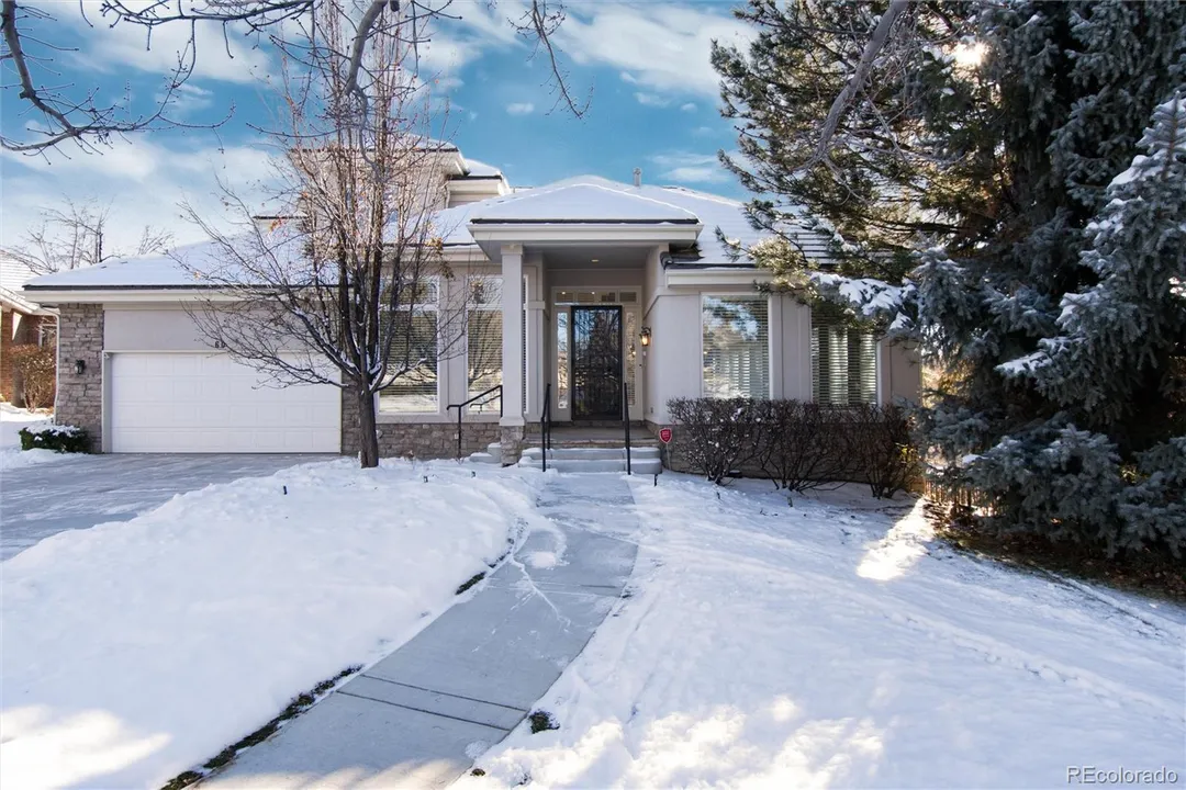 Snow-covered suburban house with a driveway, leafless tree, and large evergreen trees on the right.