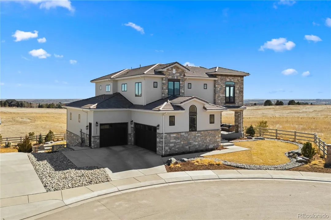 A large modern two-story house with a stone and stucco exterior, two attached garages, and a well-manicured yard surrounded by a wooden fence, set in a rural area under a blue sky with a few clouds.