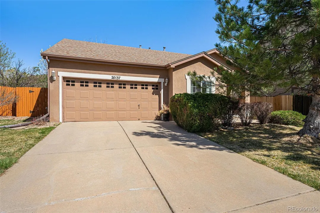 Front view of a single-family house with a driveway, beige garage door, and landscaping including bushes, trees, and a wooden fence in the background.