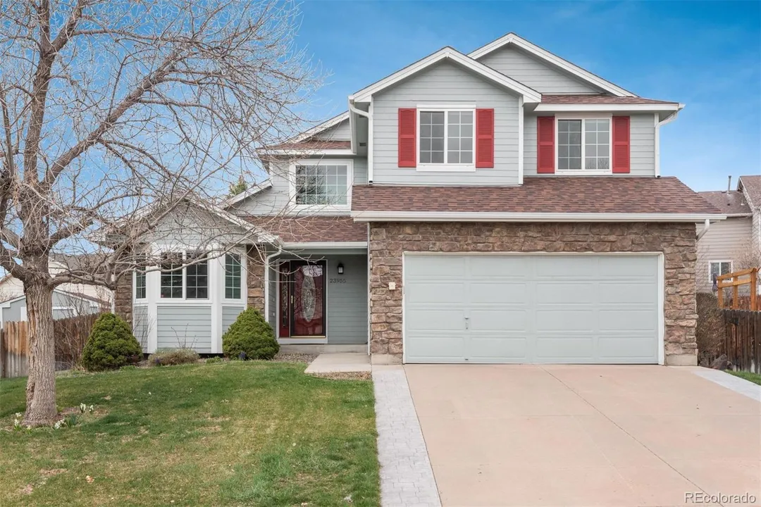 A two-story suburban house with gray siding, red shutters, and a stone facade above the garage, with a leafless tree, green lawn, and a paved driveway.
