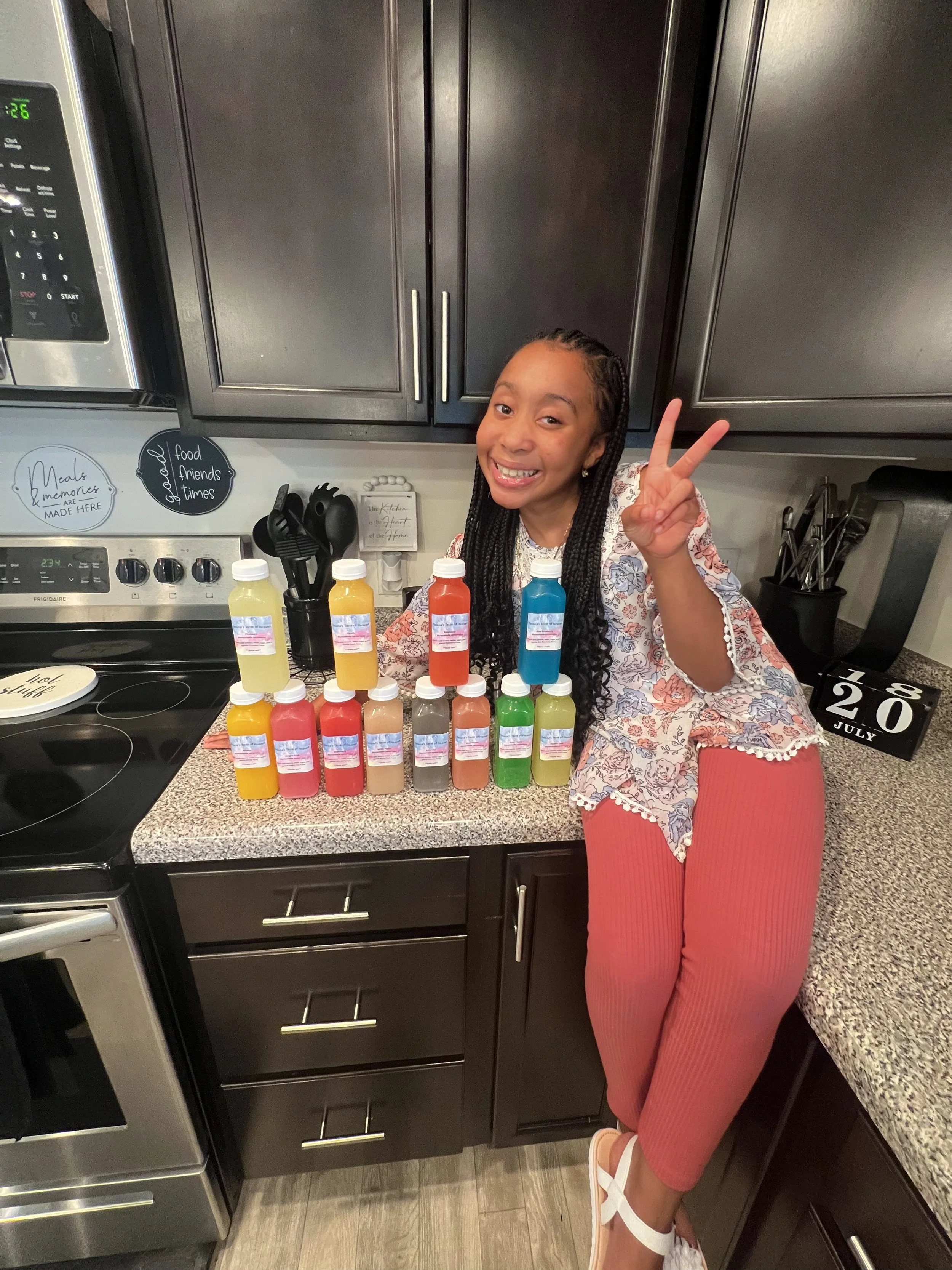 A young girl smiling and making a peace sign while standing next to a collection of colorful bottles on a kitchen countertop. Providing flavorfull homemade lemonade to your doorstep.