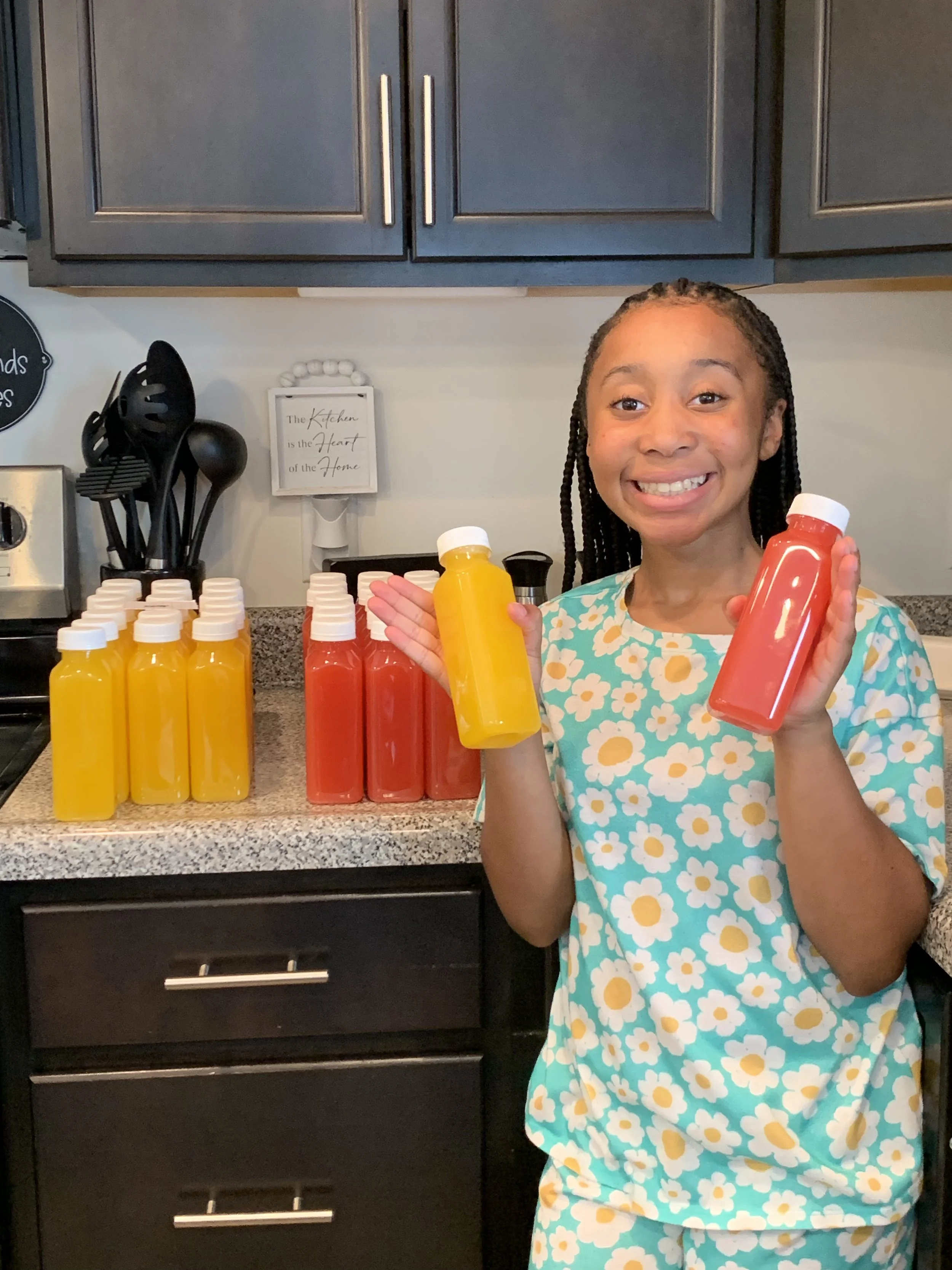 A young girl with braided hair smiling and holding a yellow and a red bottle of juice, standing in a kitchen with more bottles of juice on the counter. Providing flavorfull homemade lemonade to your doorstep.