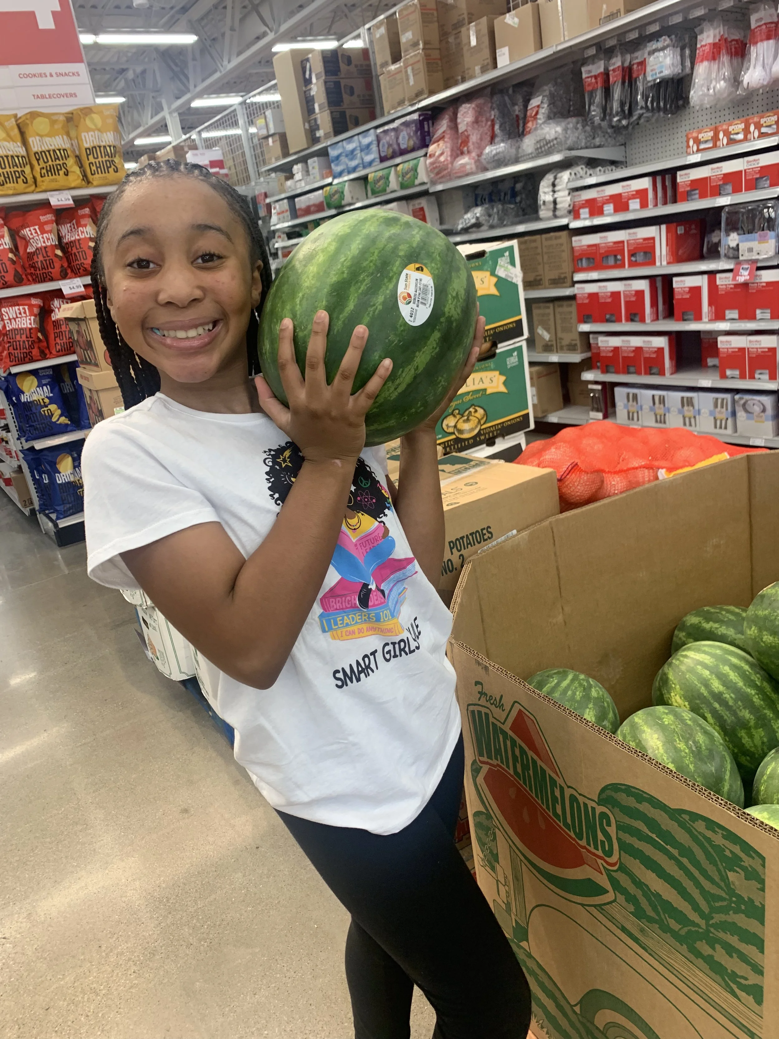 A young girl holding a watermelon, green watermelon, at the grocery store. Providing flavorfull homemade lemonade to your doorstep.