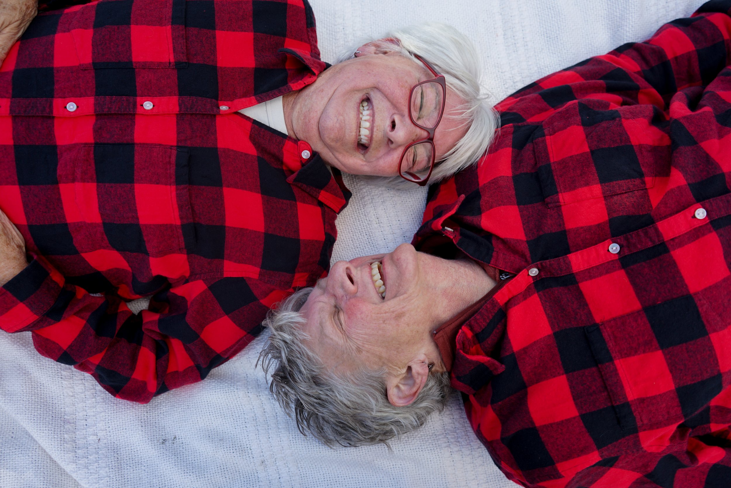 Two elderly women lying down back-to-back, smiling and wearing matching red and black plaid shirts.
