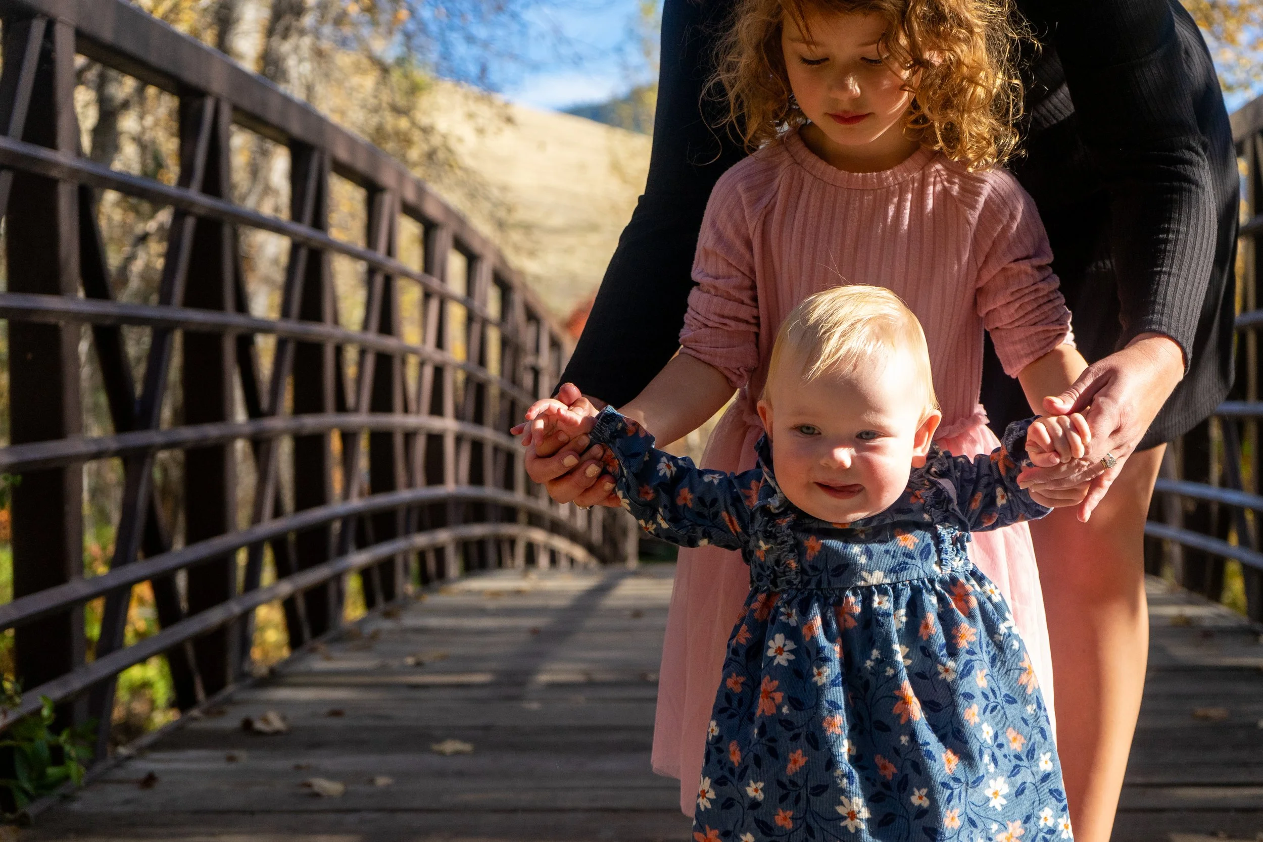 A young girl with curly hair in a pink dress supports a younger girl with blond hair and a blue floral dress on a wooden bridge outdoors during autumn.