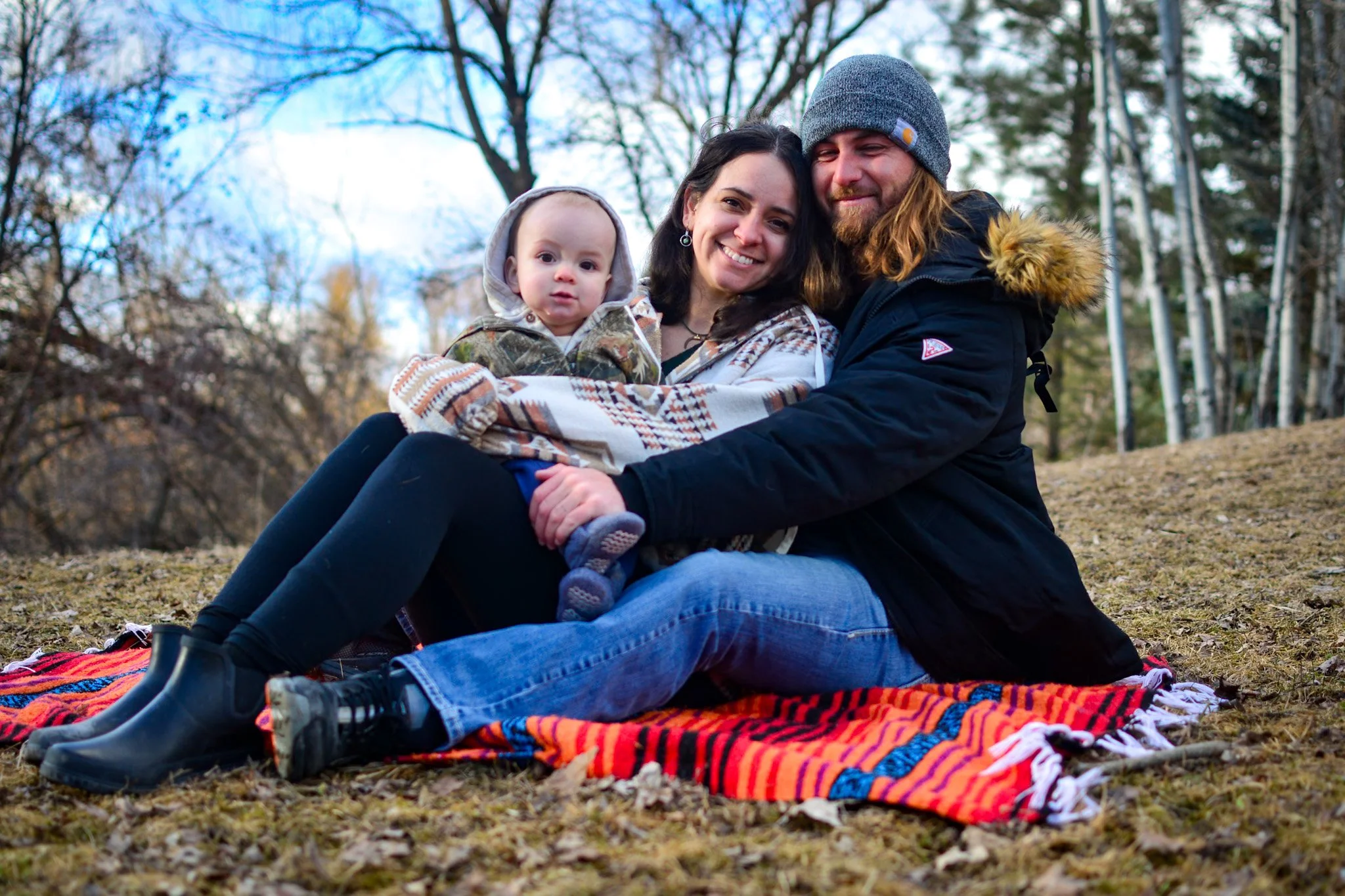 A family of three sitting on a colorful blanket outdoors in a park during fall, with trees in the background. The woman is holding a child while the man sits beside them, all smiling.
