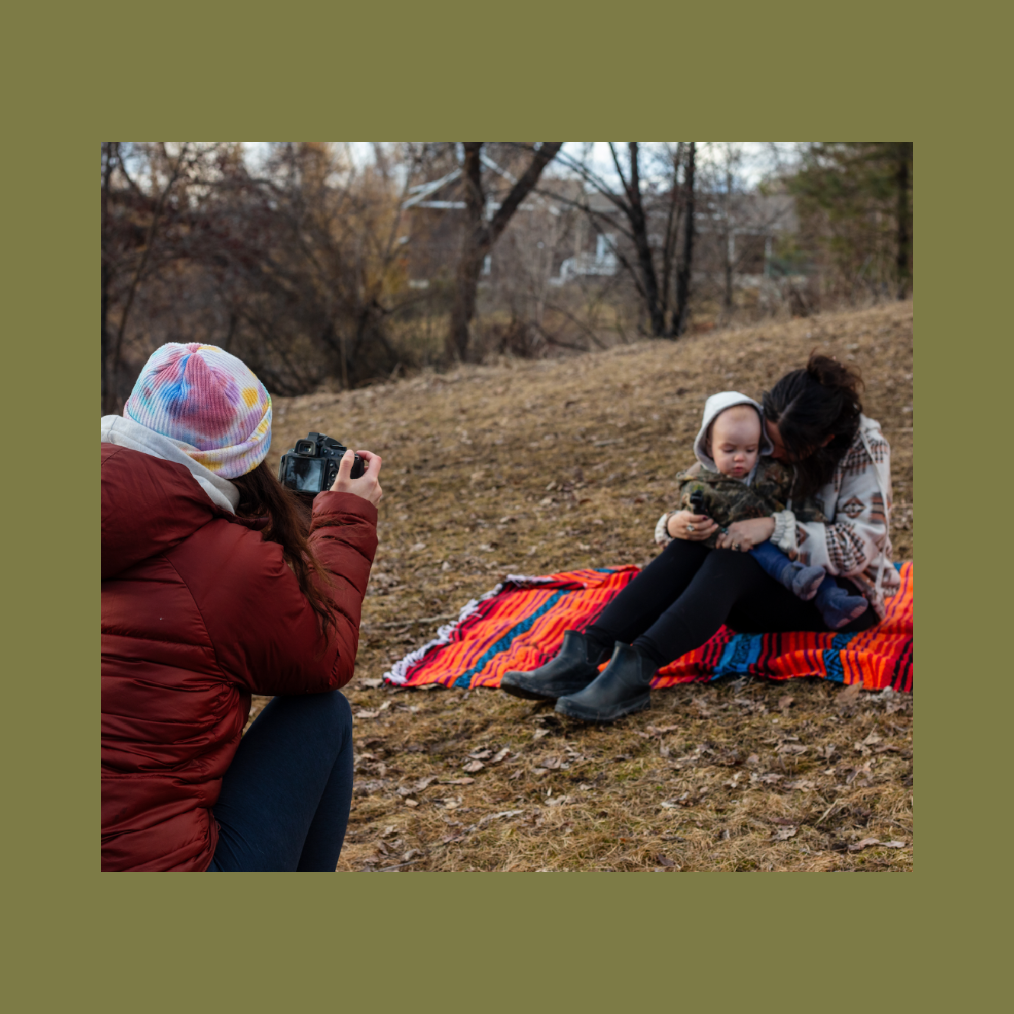 People enjoying outdoor fall day, sitting on colorful blanket, children and adult, one child wearing tie-dye hat, another person taking photo.