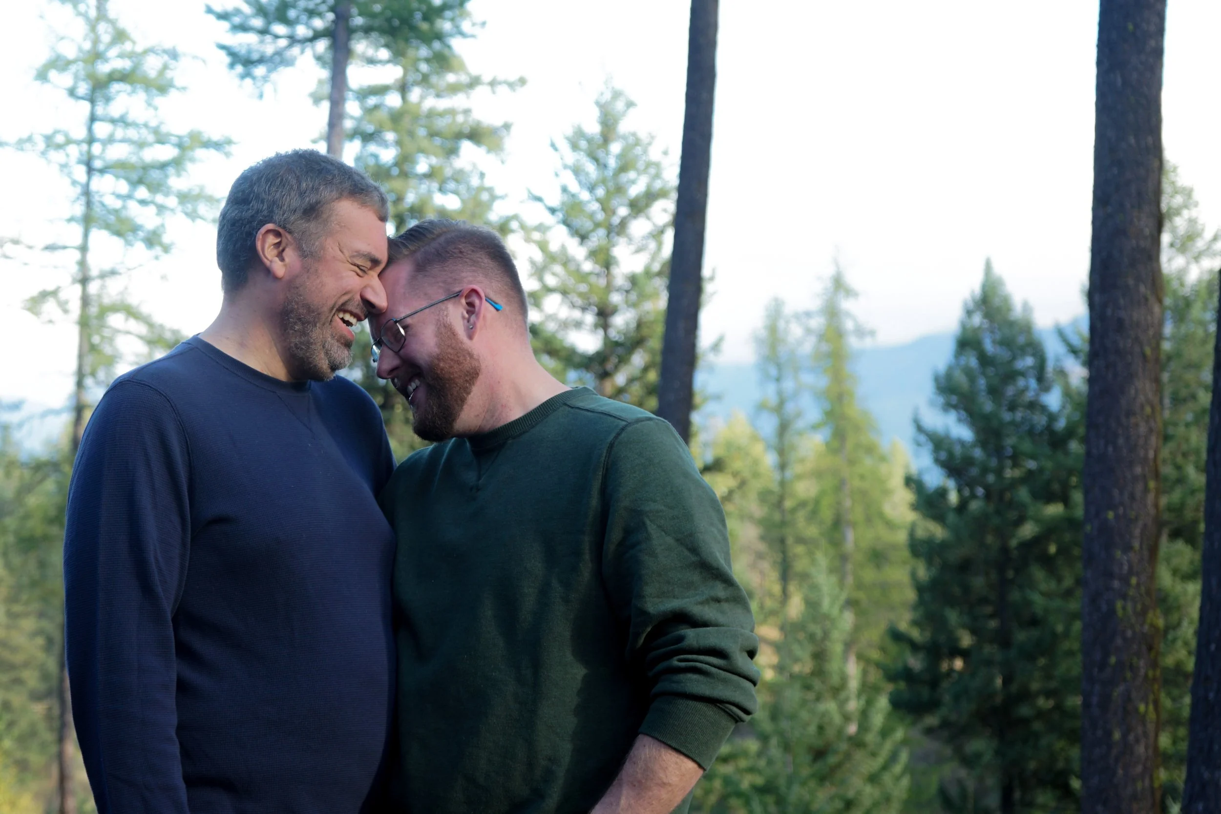 Two men smiling and sharing a close, joyful moment outdoors in a forest with tall trees and mountains in the background.