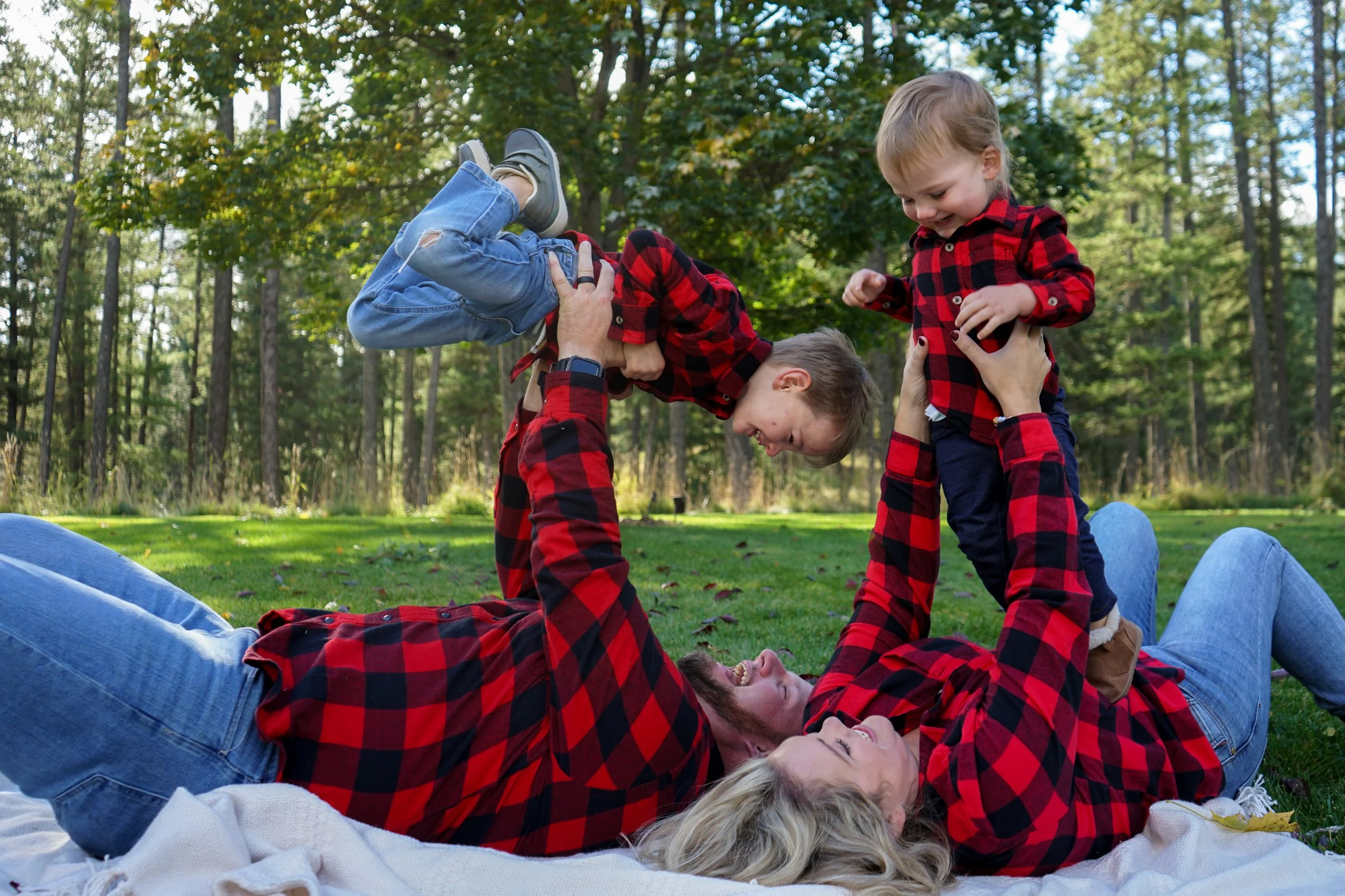 A family of four, all wearing matching red and black plaid shirts, playing on a blanket in a grassy park with trees in the background. The father lifts a young boy into the air while the mother and another child watch and smile.