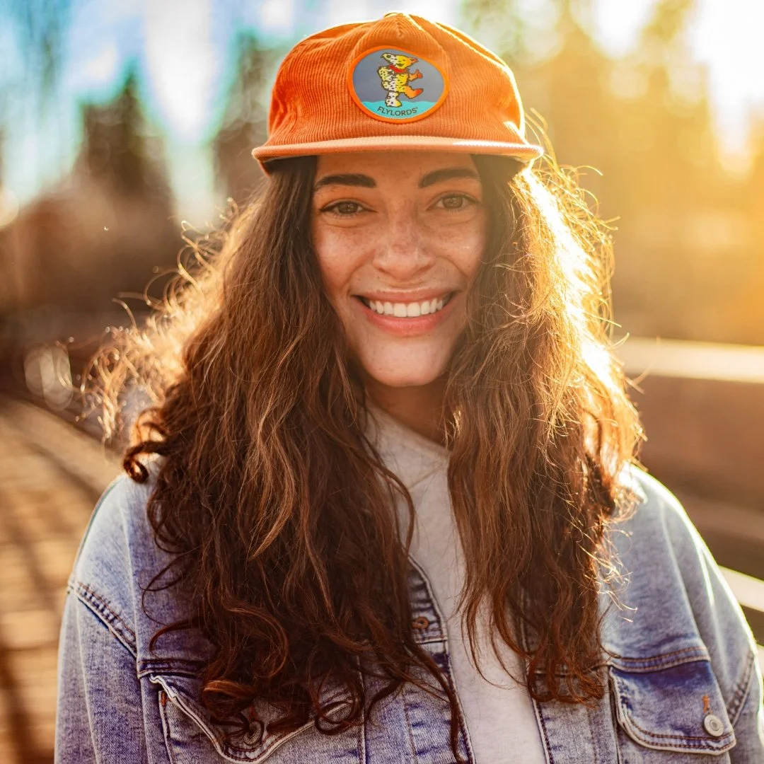 A woman with long curly hair smiling outdoors in warm sunlight, wearing an orange cap with a patch and a denim jacket.
