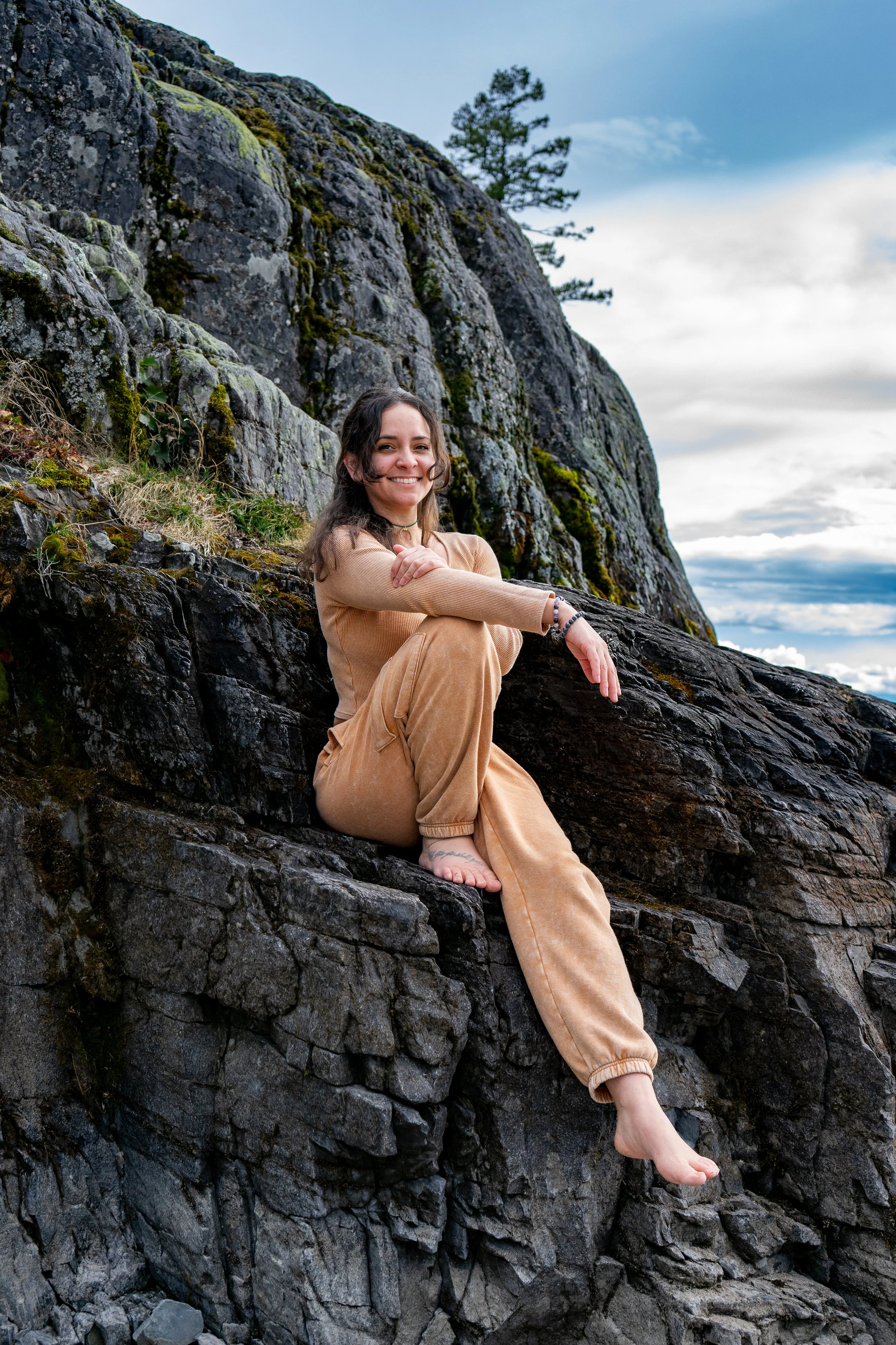 A woman sitting on a large rock formation outdoors with a hillside covered in moss and small plants, looking at the camera with a smile, wearing a tan hoodie and matching pants, foot resting on a lower rock, with a cloudy sky and a tree in the backgr