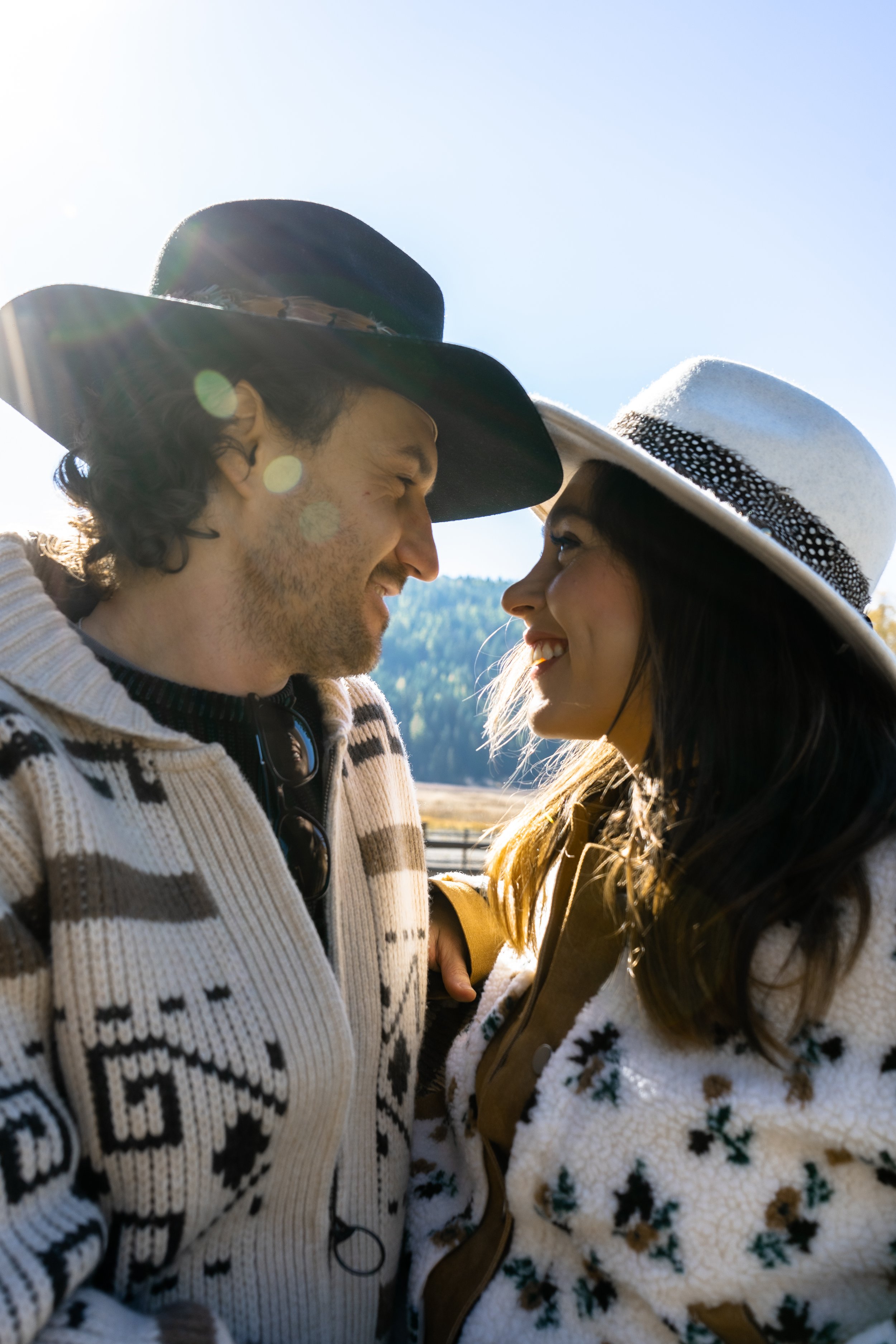 A man and woman, both smiling and close to each other, wearing wide-brimmed hats and winter sweaters, standing outdoors on a sunny day with a mountain and trees in the background.
