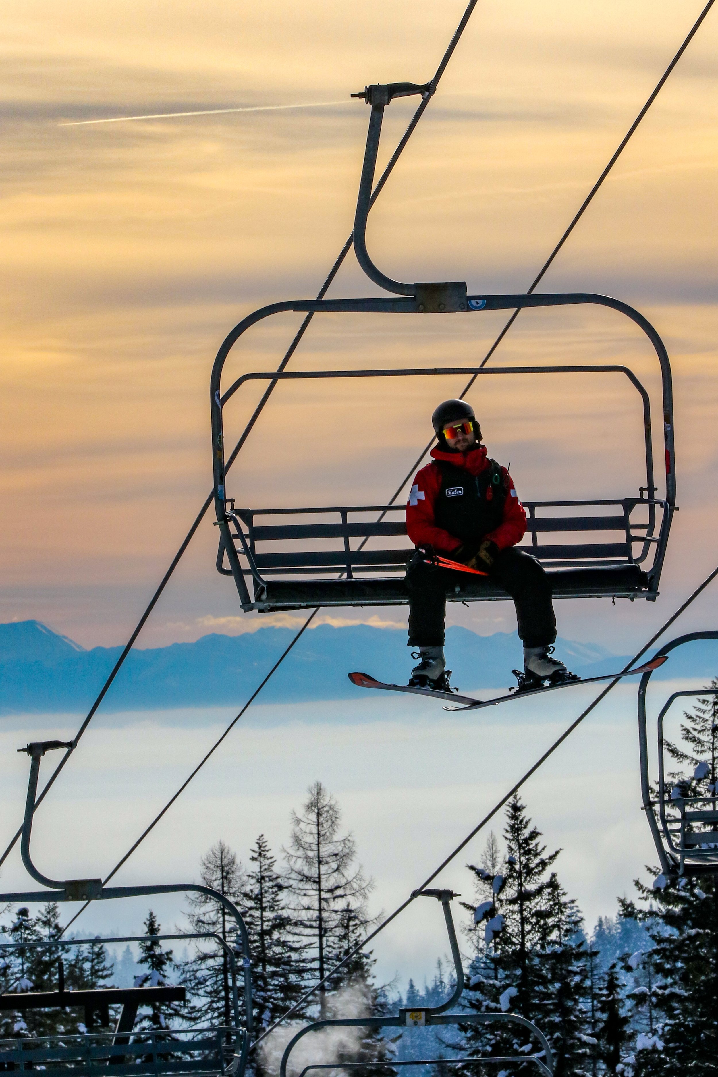 Person wearing a black helmet, red and black ski jacket, and ski goggles sitting on a ski lift with snowy mountain and forest in the background during sunset.