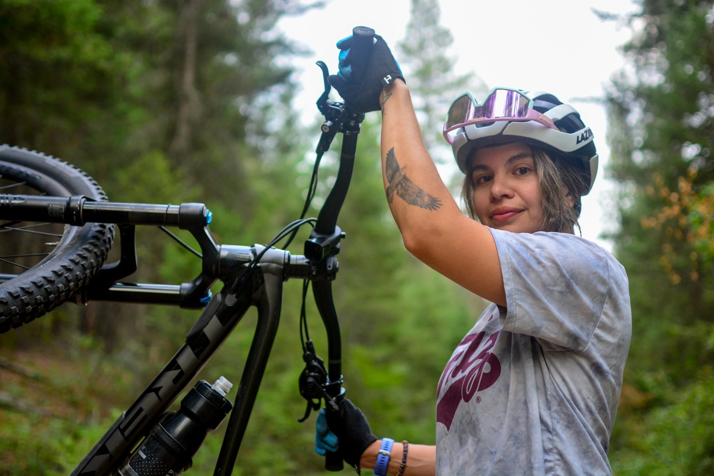 A woman in a helmet and gloves is fixing a mountain bike in a wooded outdoor area.