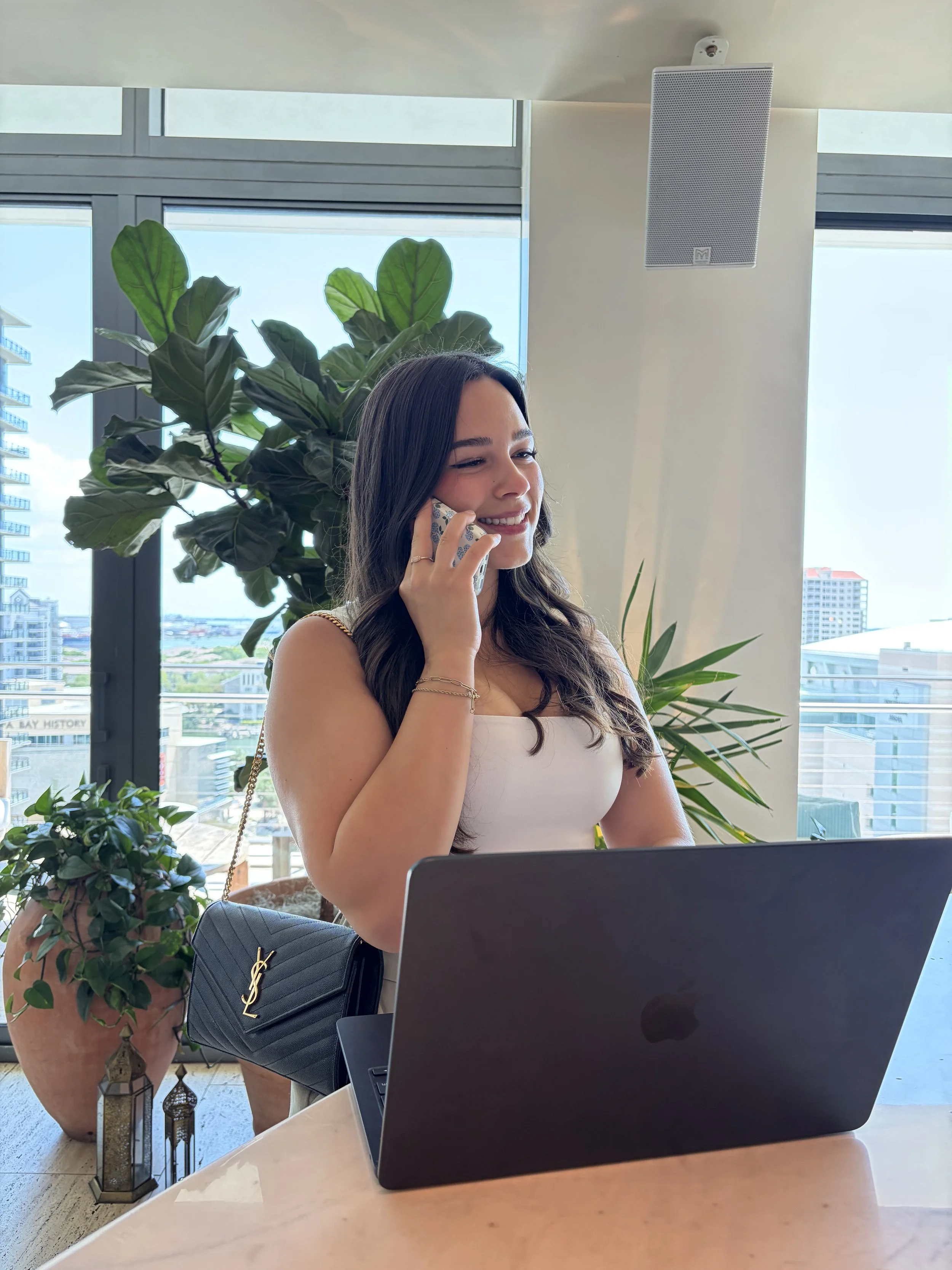 A woman talking on her cellphone in an office with large windows, a laptop, and green plants.