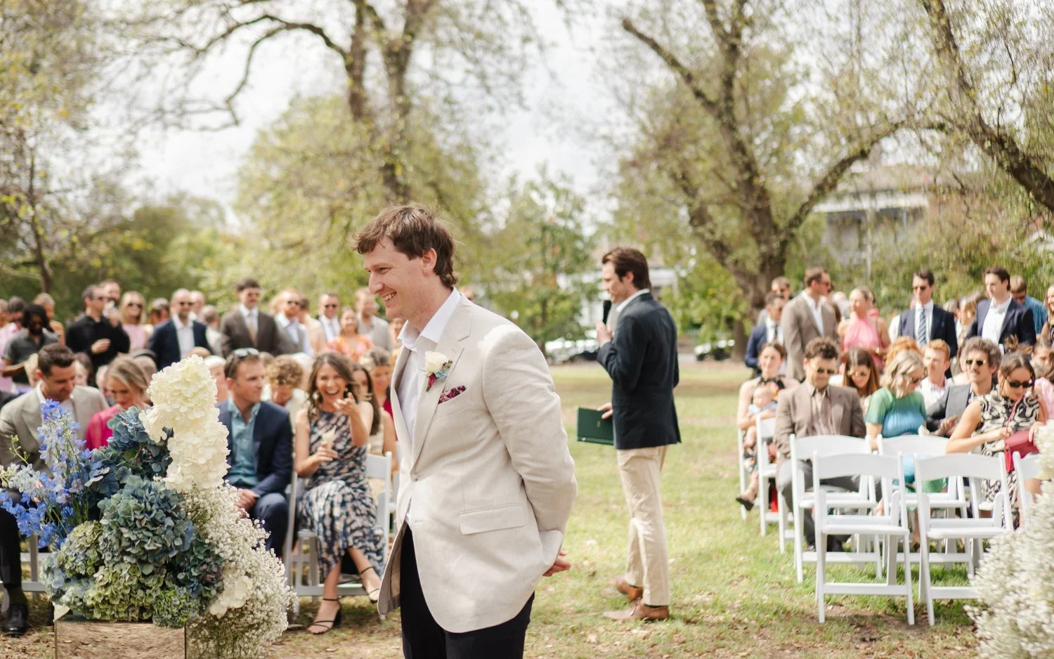 A groom in a cream-colored suit smiling at his wedding ceremony outdoors, with guests seated and standing under trees in the background.