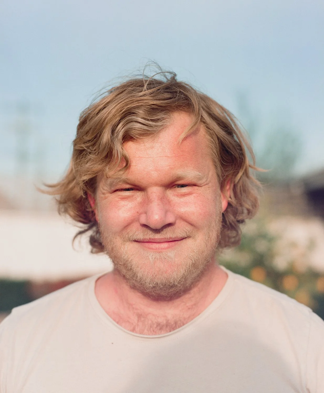 Close-up of a man with blond curly hair, light beard, and a slight smile, wearing a light-colored t-shirt outdoors.