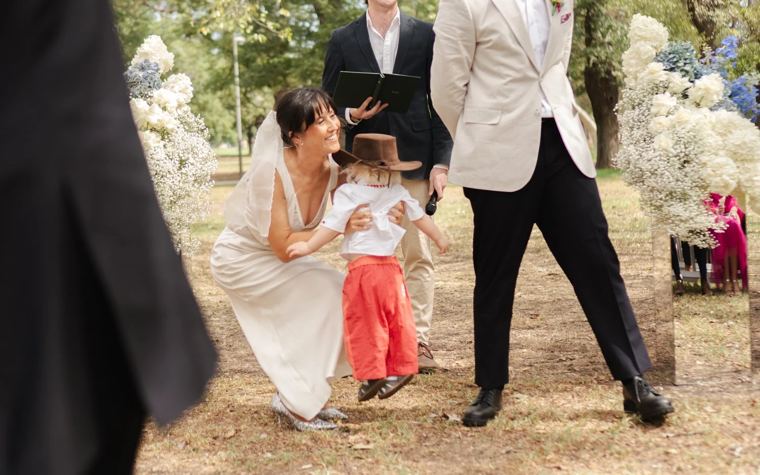 A woman in a white dress smiling and kneeling to hold a young child wearing a cowboy hat, white shirt, and red pants at an outdoor wedding ceremony. Other guests are present, and there are white flowers decorated around.