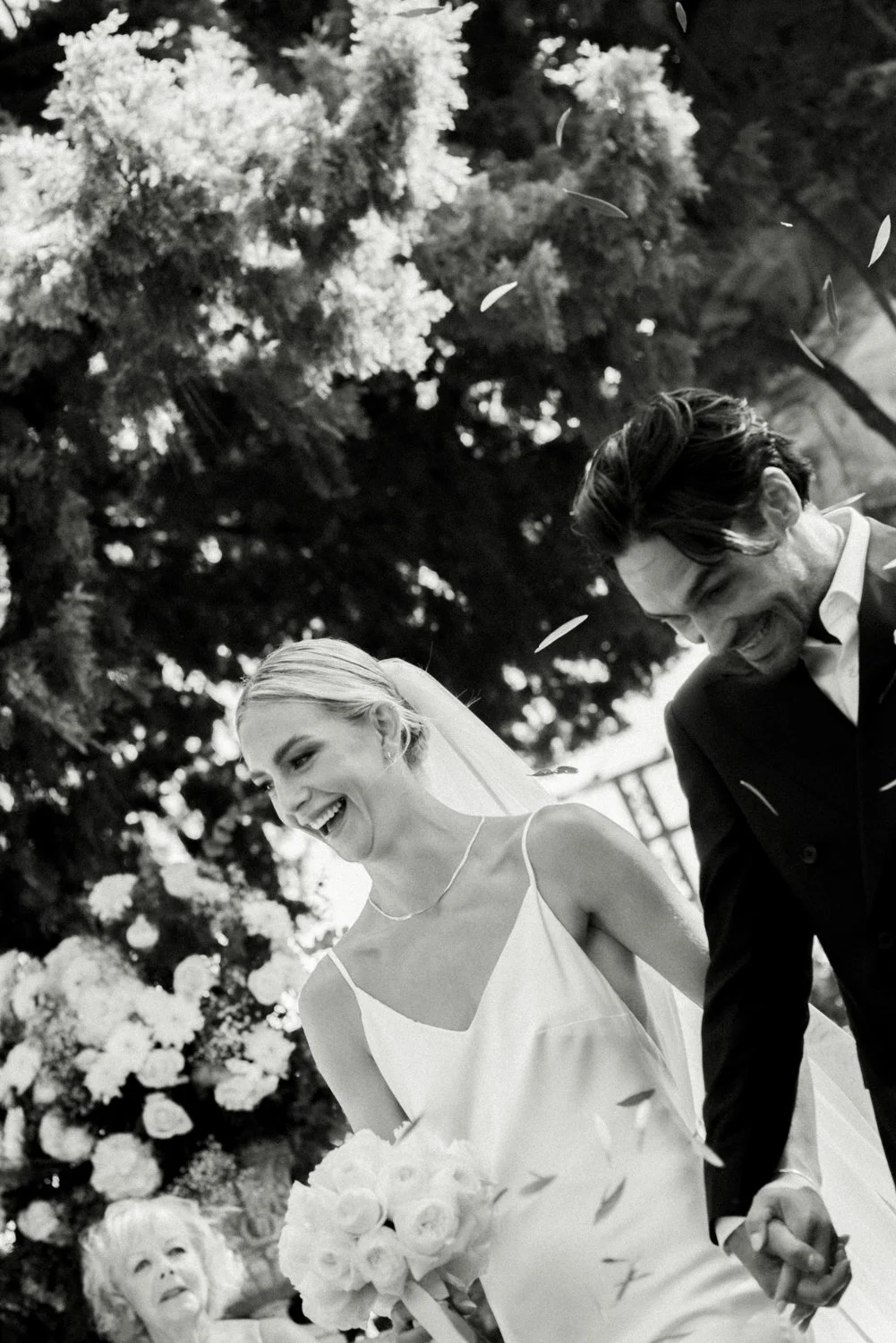 Black and white photo of a smiling bride in a white dress holding a bouquet, and a groom in a dark suit, holding hands and smiling during a wedding ceremony outdoors with a woman and trees in the background.