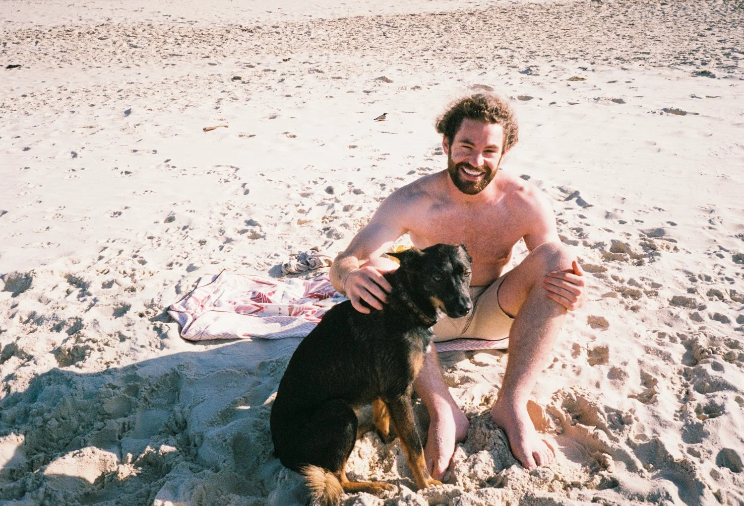 A man with a beard sitting on the sandy beach, smiling, with a black and brown dog sitting in front of him.
