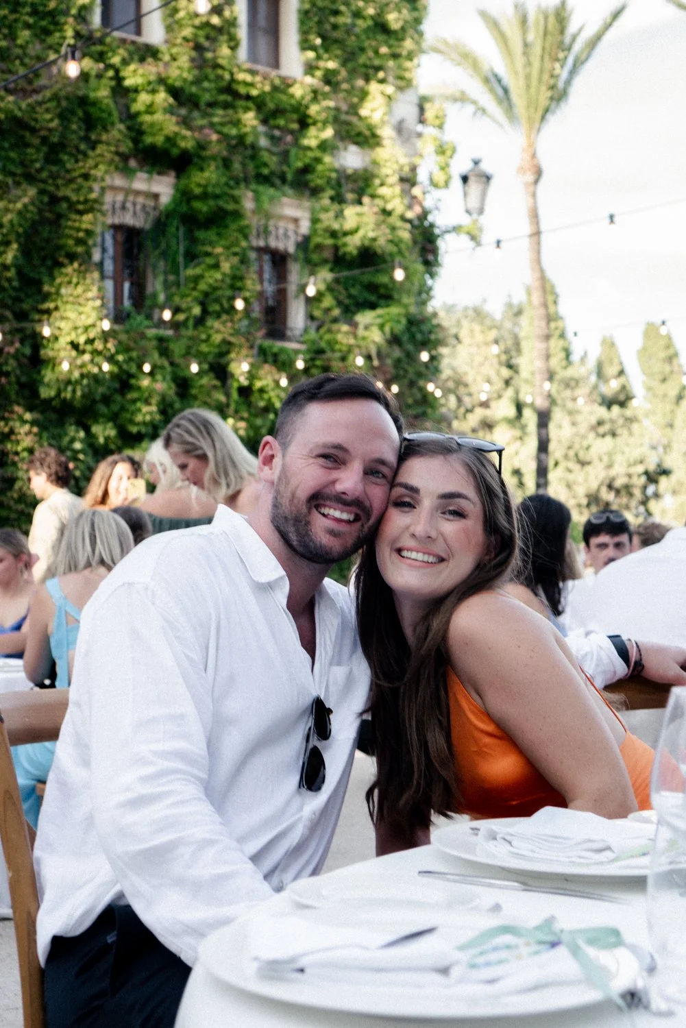 A smiling man and woman sitting at an outdoor event, with a green ivy-covered building and string lights in the background.
