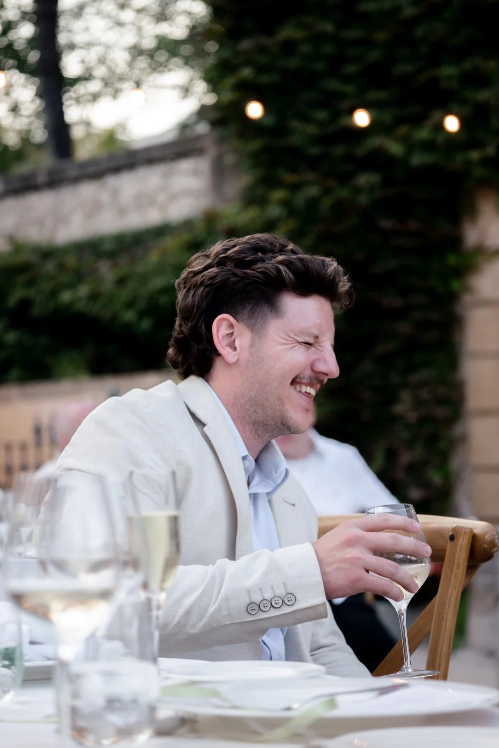 A man with curly brown hair wearing a light-colored suit smiling and laughing while holding a glass of white wine at an outdoor gathering.
