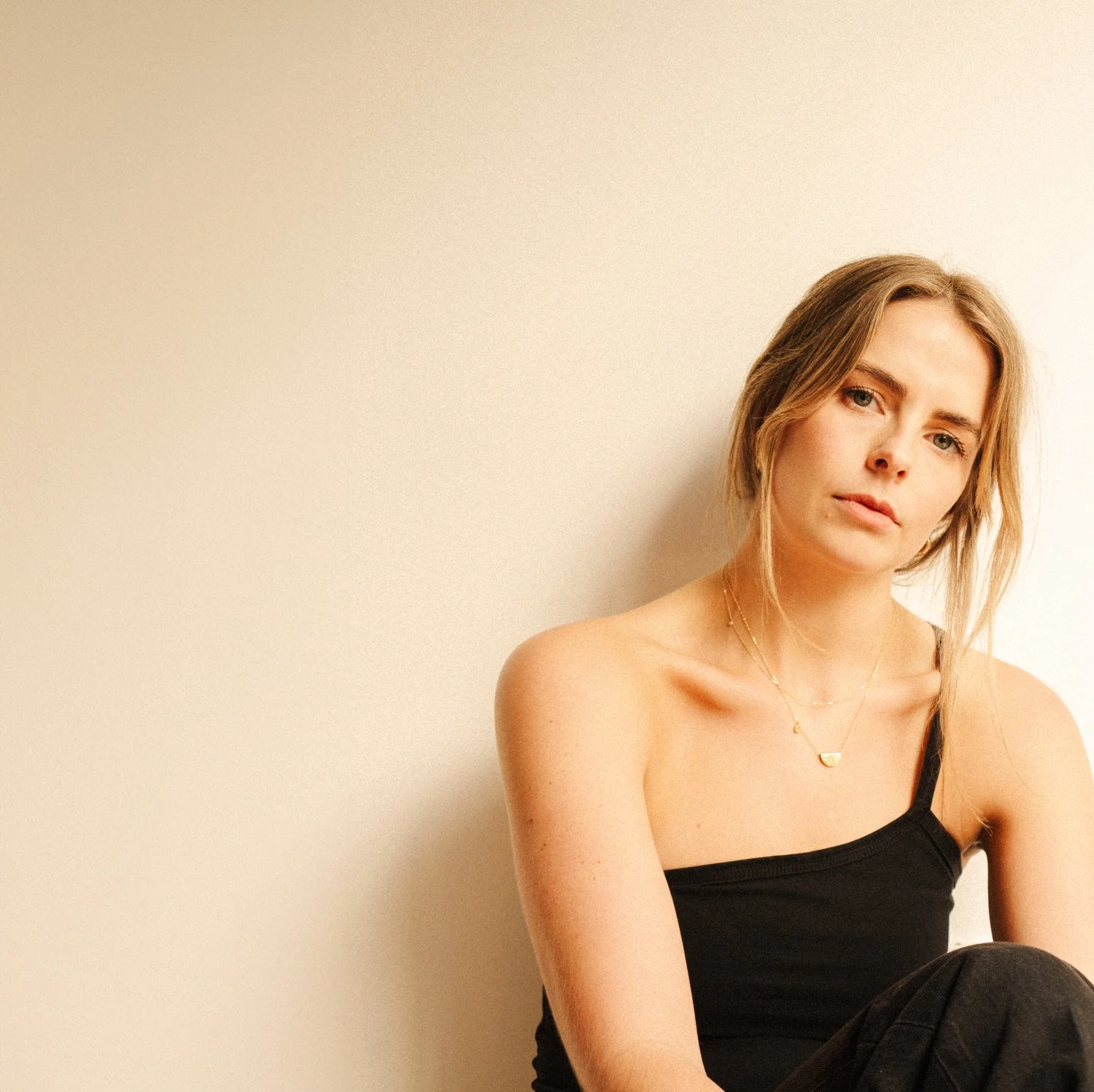 A young woman with blonde hair and fair skin wearing a black spaghetti strap top and gold jewelry, sitting against a plain beige wall