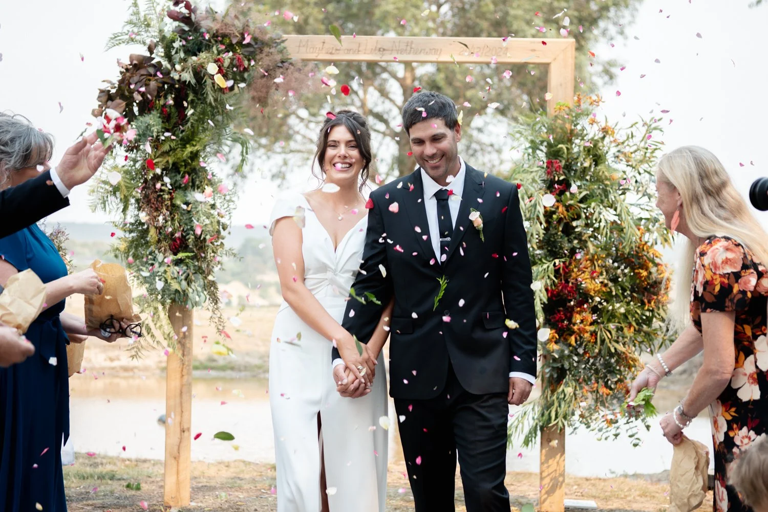 A couple in wedding attire holding hands and smiling as guests throw flower petals at an outdoor wedding ceremony.