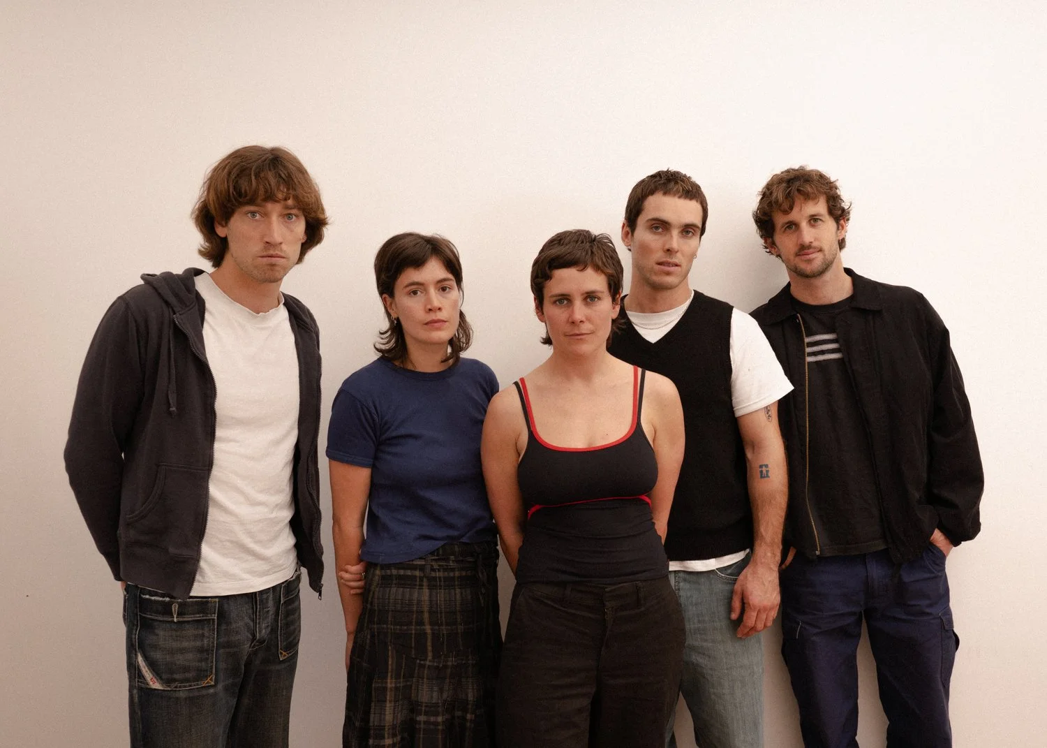 Group of five young adults standing against a plain white wall, looking at the camera with neutral expressions.