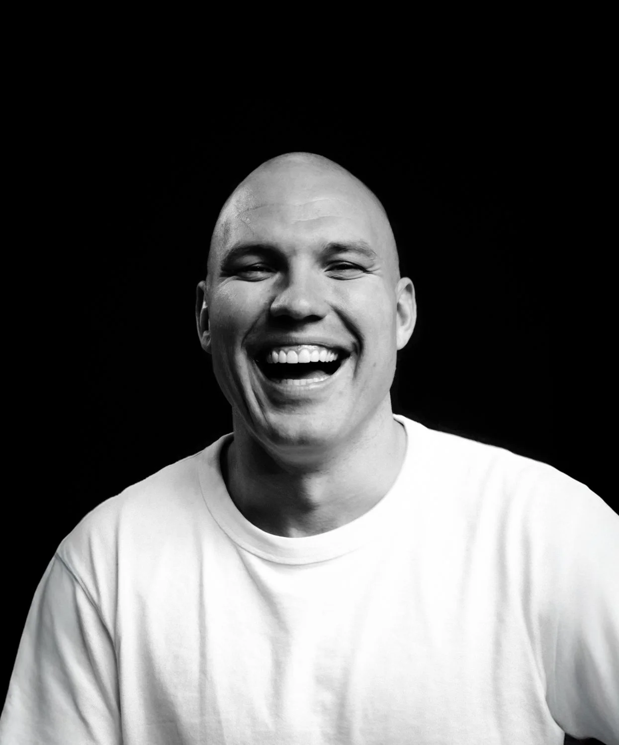 Black and white photo of a smiling man with a bald head, wearing a white t-shirt, on a black background.