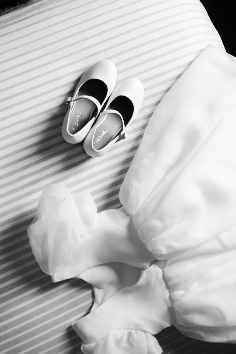 A pair of white shoes placed on a striped bedspread next to a pillow and a white dress.