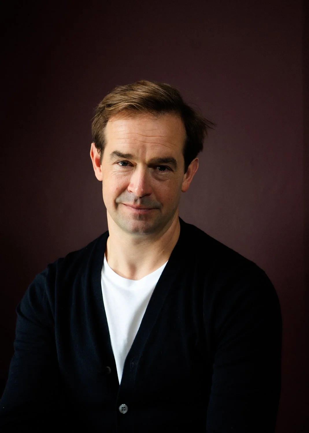 Portrait of a smiling man with short brown hair, wearing a black cardigan over a white shirt, set against a dark background.