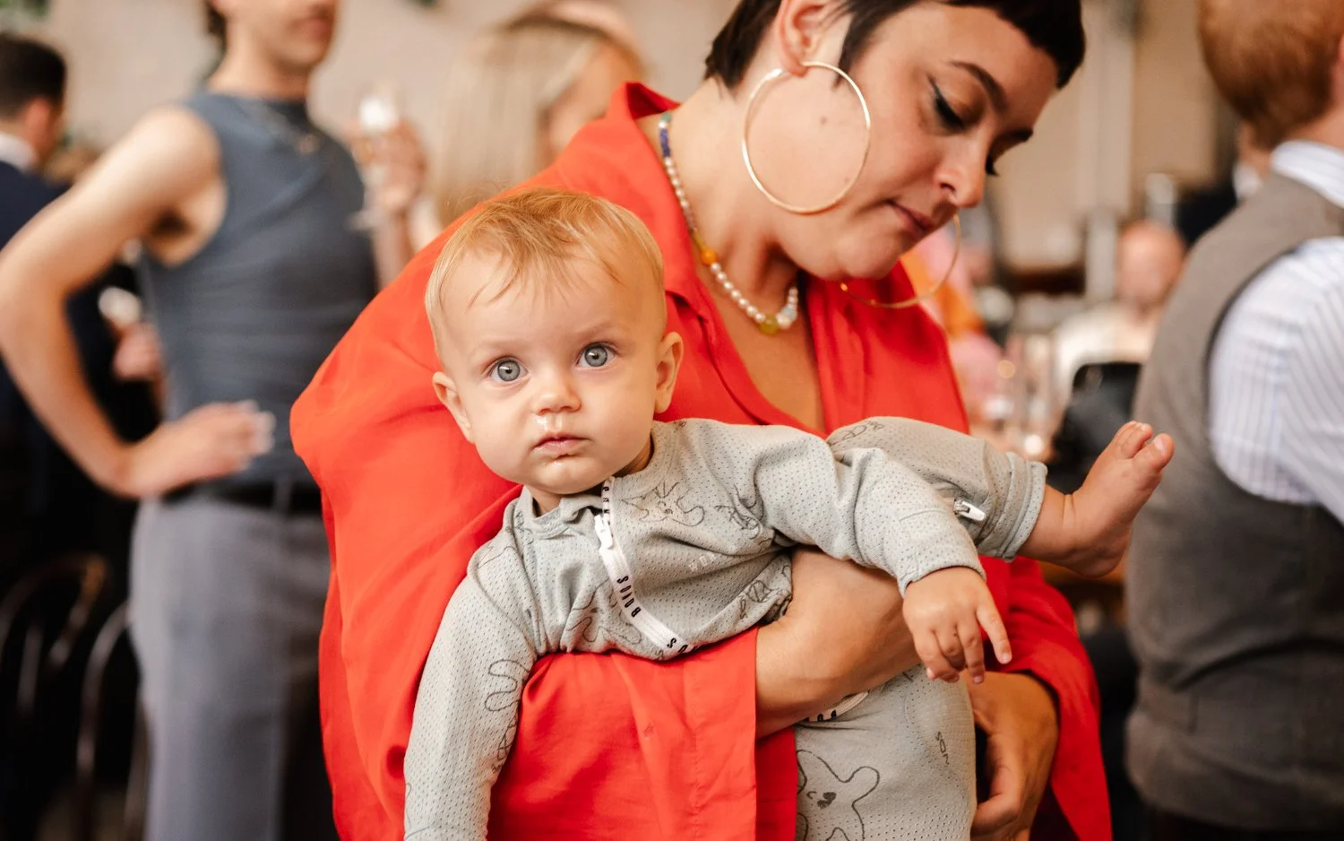 A woman holding a baby at a social event or gathering, with other people in the background.