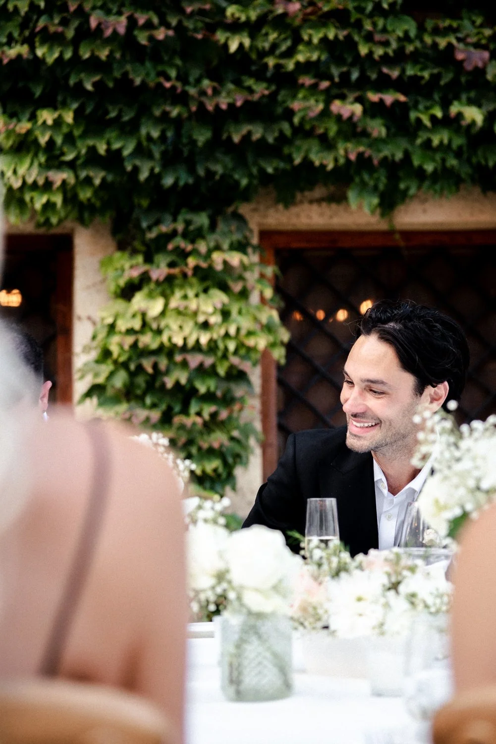 A man in a black suit and white shirt smiling at a wedding reception table decorated with white flowers.